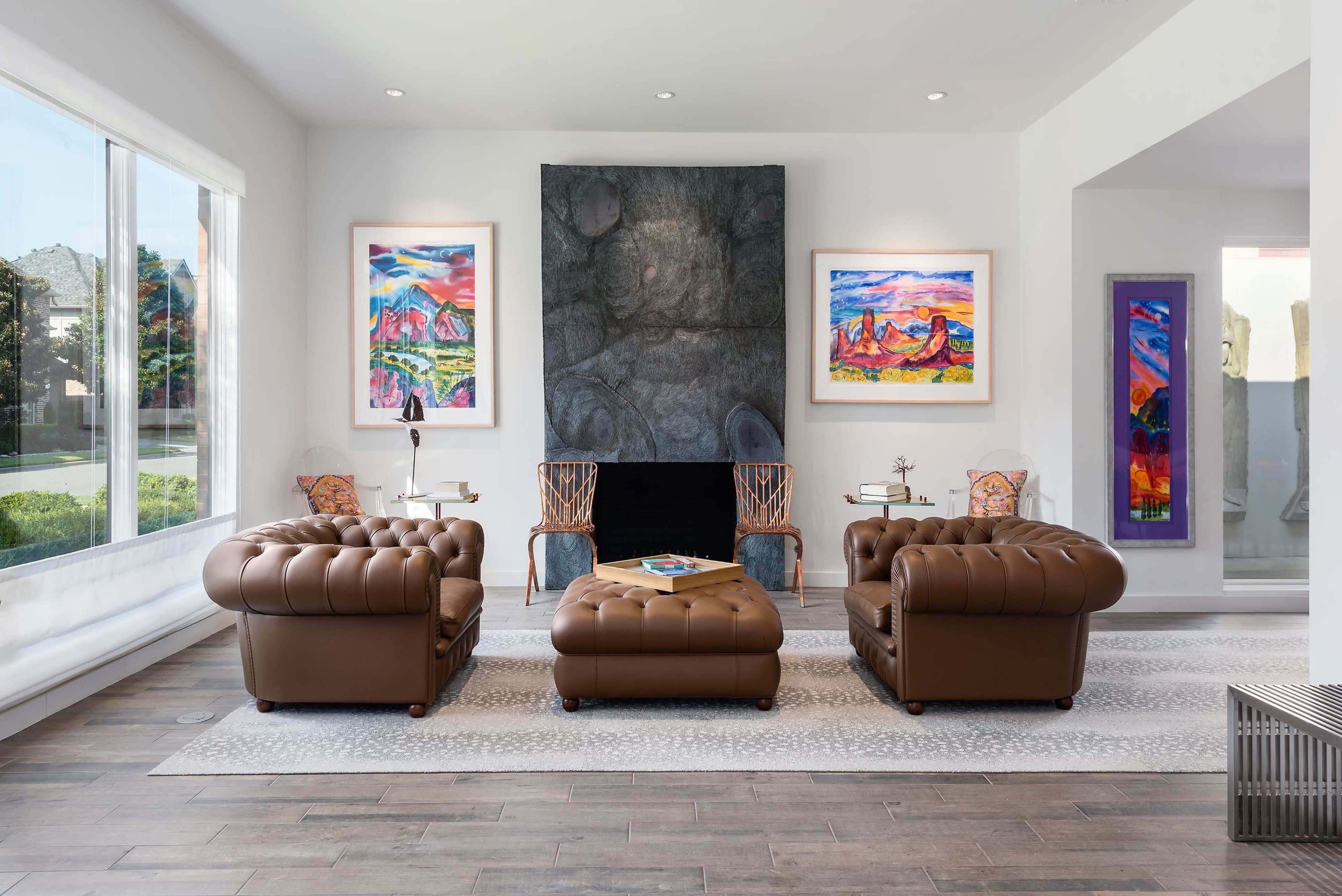 A modern living room featuring two brown tufted armchairs, a coffee table, colorful artwork on the walls, and a stone fireplace.