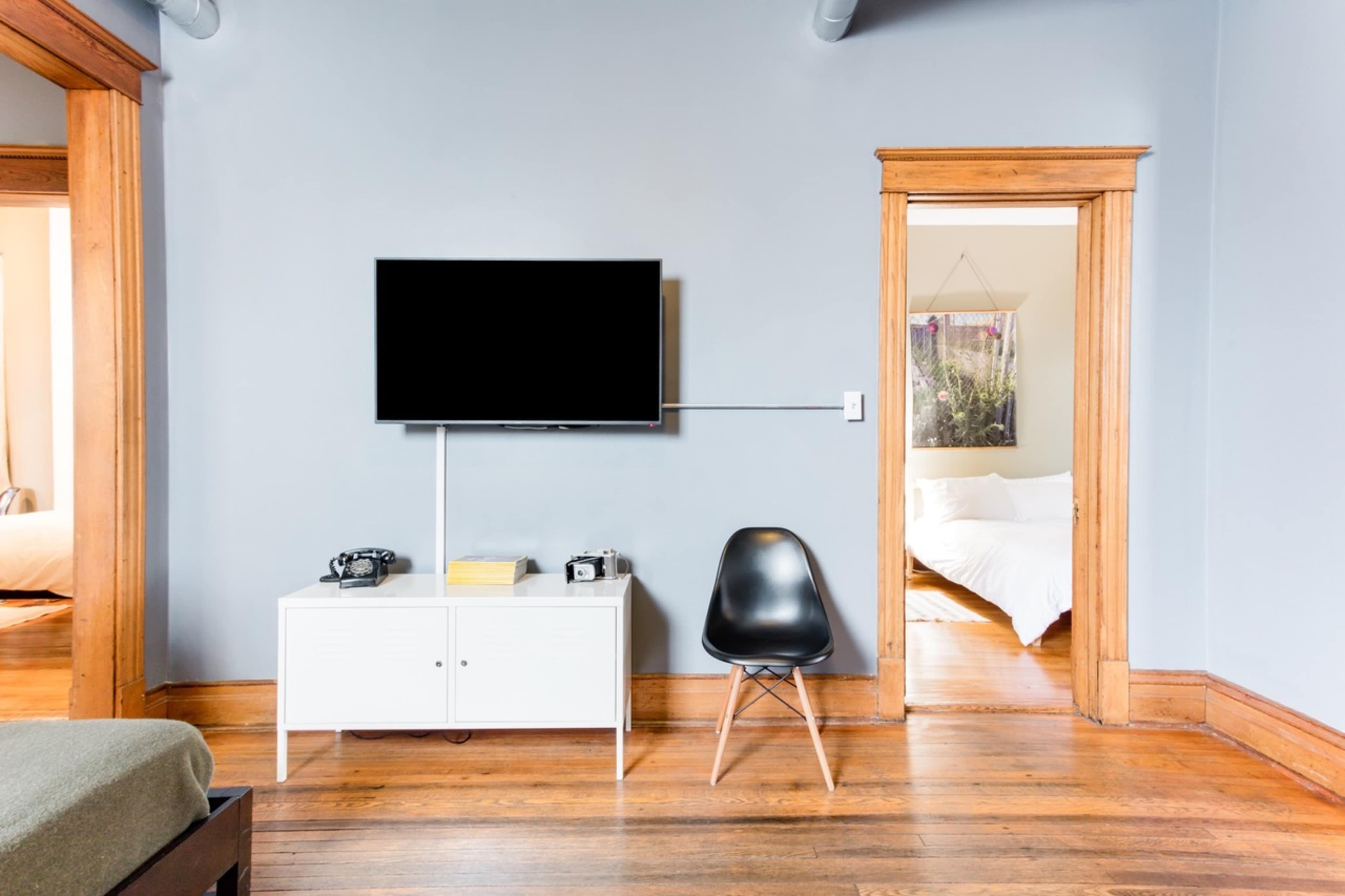 A living space features a wall-mounted television, a white media console with cameras and books, and a black chair, while a doorway leads to a bedroom with a white bed.