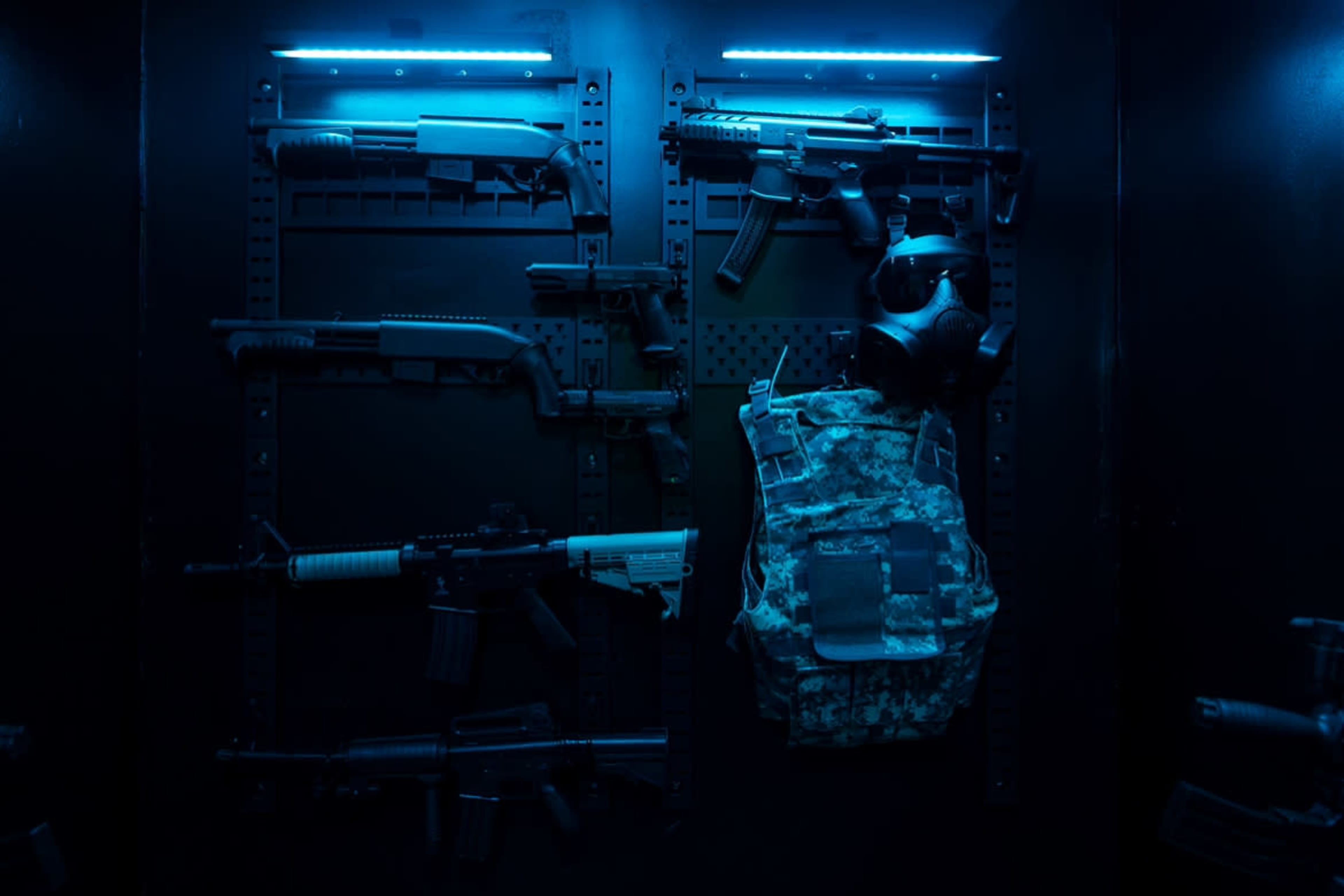 A display of various firearms and tactical gear, including a gas mask and a military vest, illuminated by blue lighting.