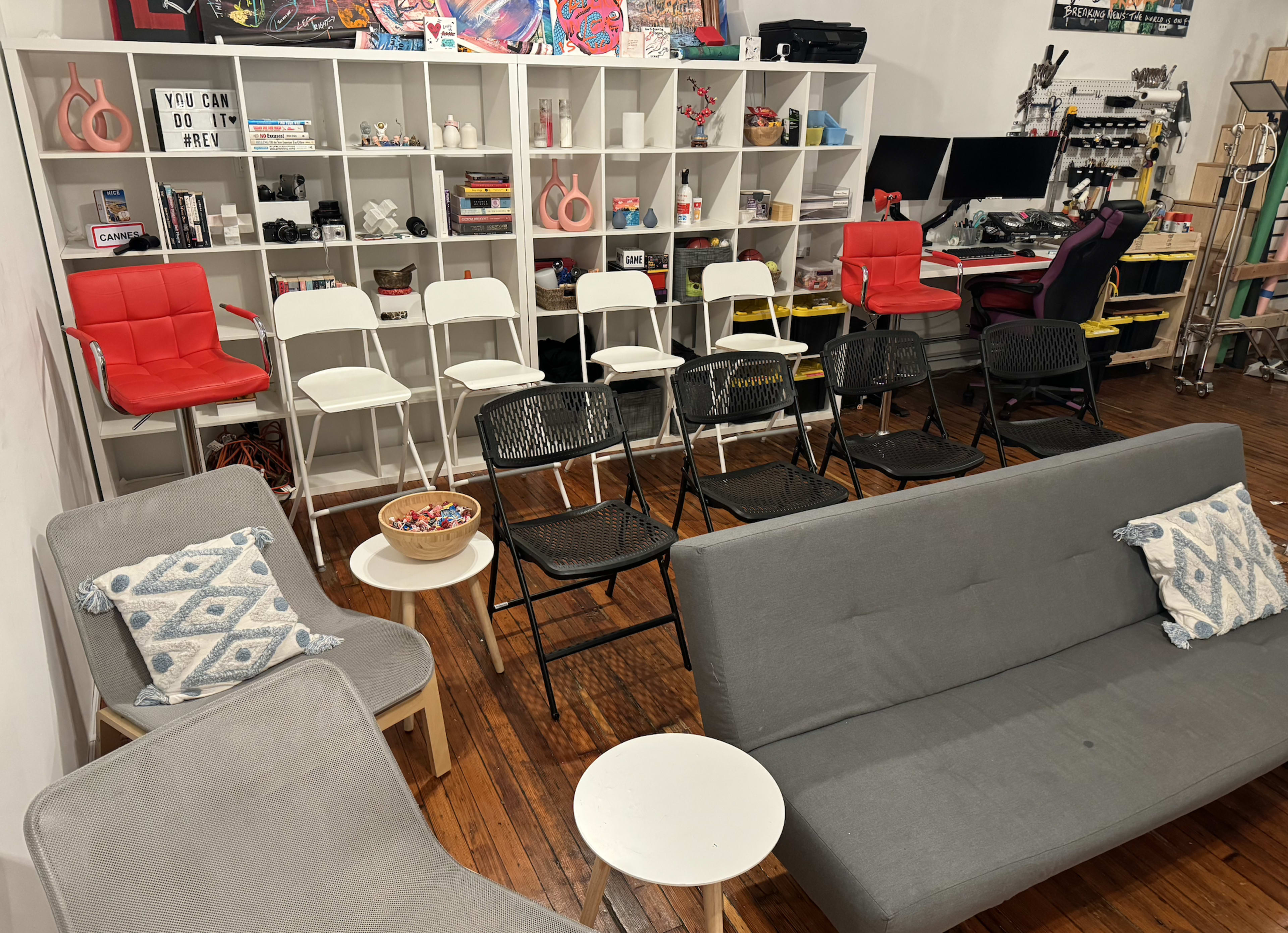 The image shows a cozy workspace with gray sofas, a white side table, a row of chairs in various colors, and a shelf filled with decorative items and equipment.