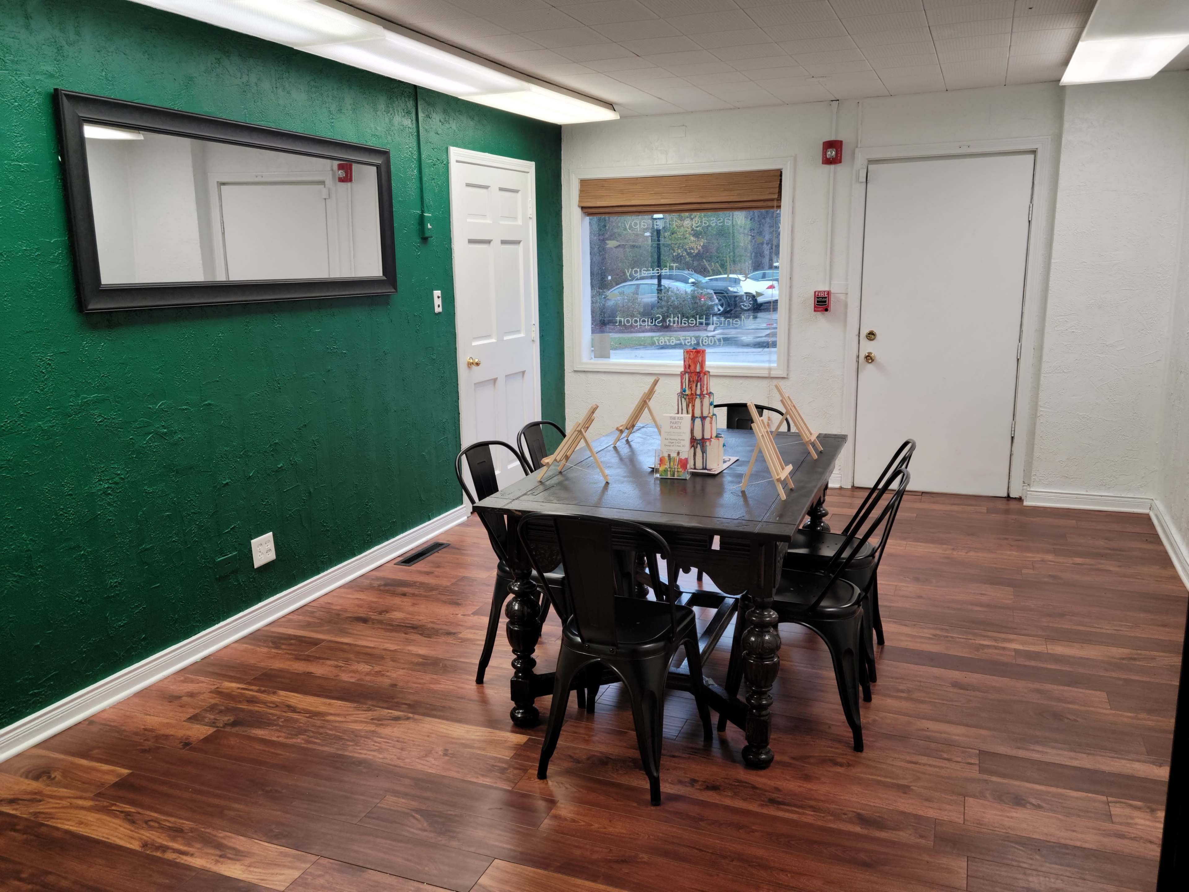 A room with a green accent wall, a large black dining table surrounded by black chairs, and a window with blinds letting in natural light.