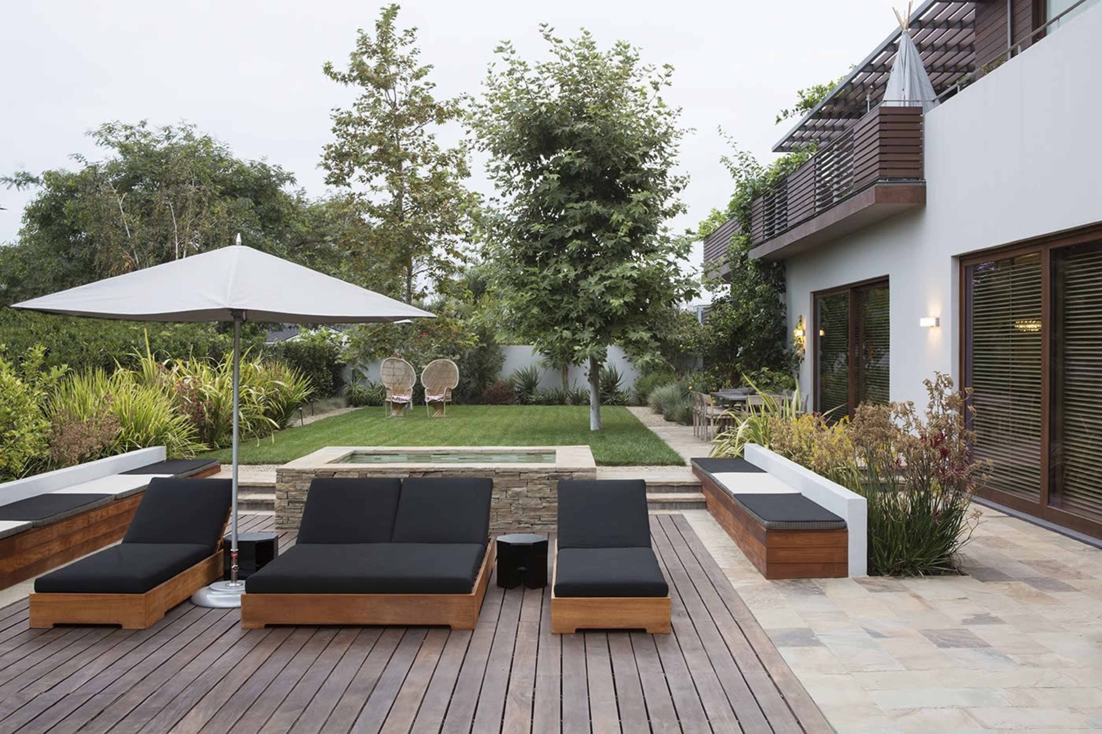 A modern outdoor patio with wooden decking, lounge chairs, an umbrella, and a landscaped garden featuring a small water feature.