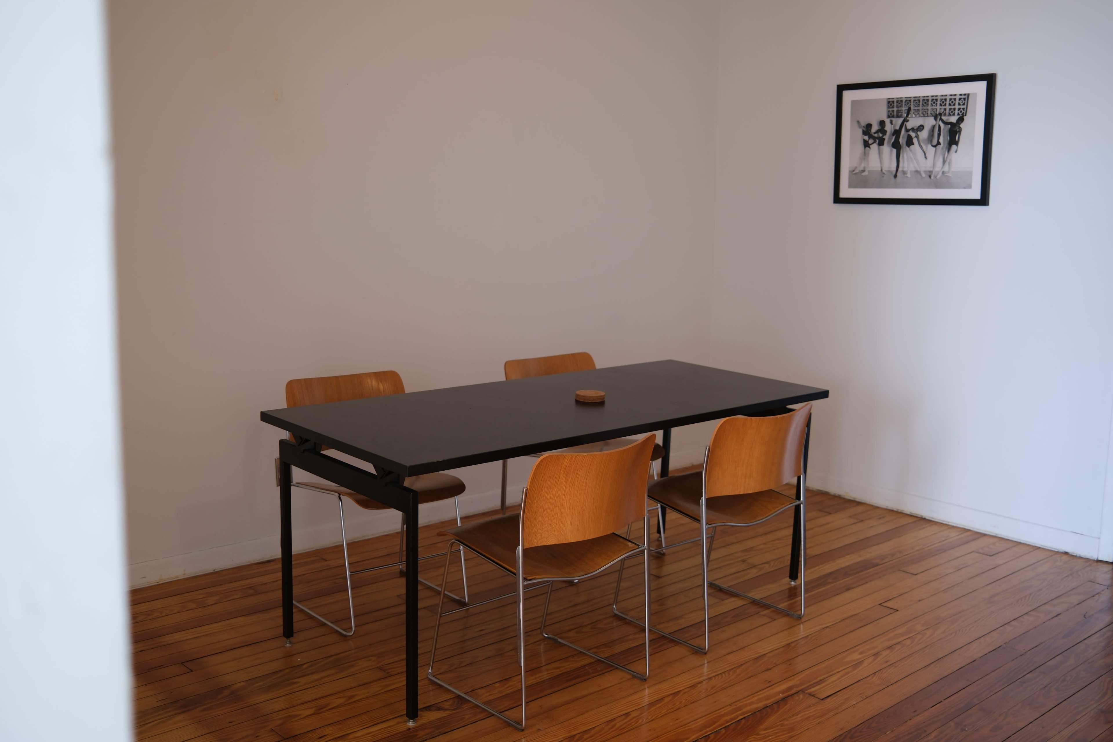A minimalist dining area features a black table surrounded by four wooden chairs and a wall-mounted black-and-white photograph.