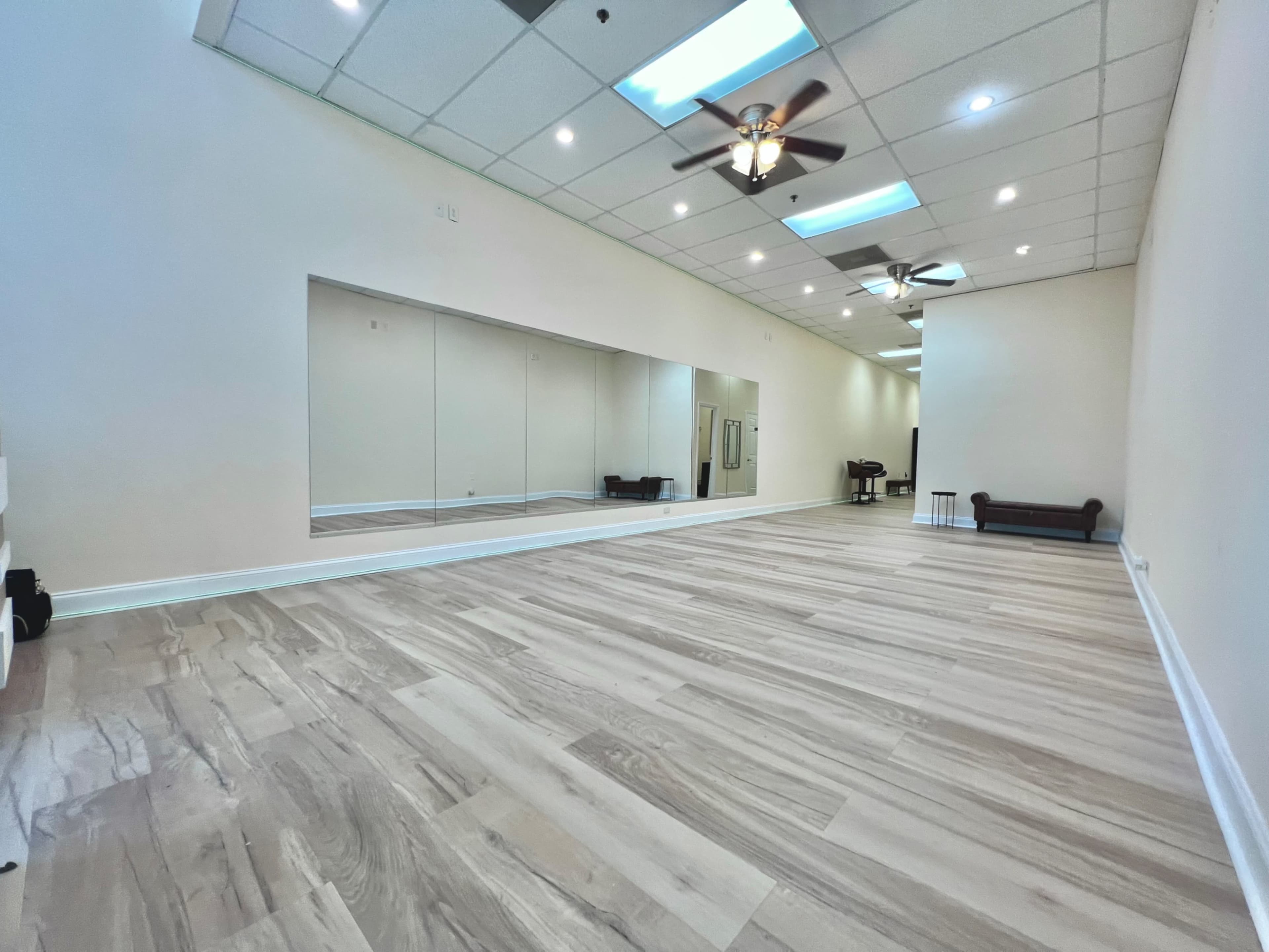 The image shows a spacious, empty room with light-colored flooring, large mirrors on one wall, and a ceiling with multiple light fixtures and ceiling fans.