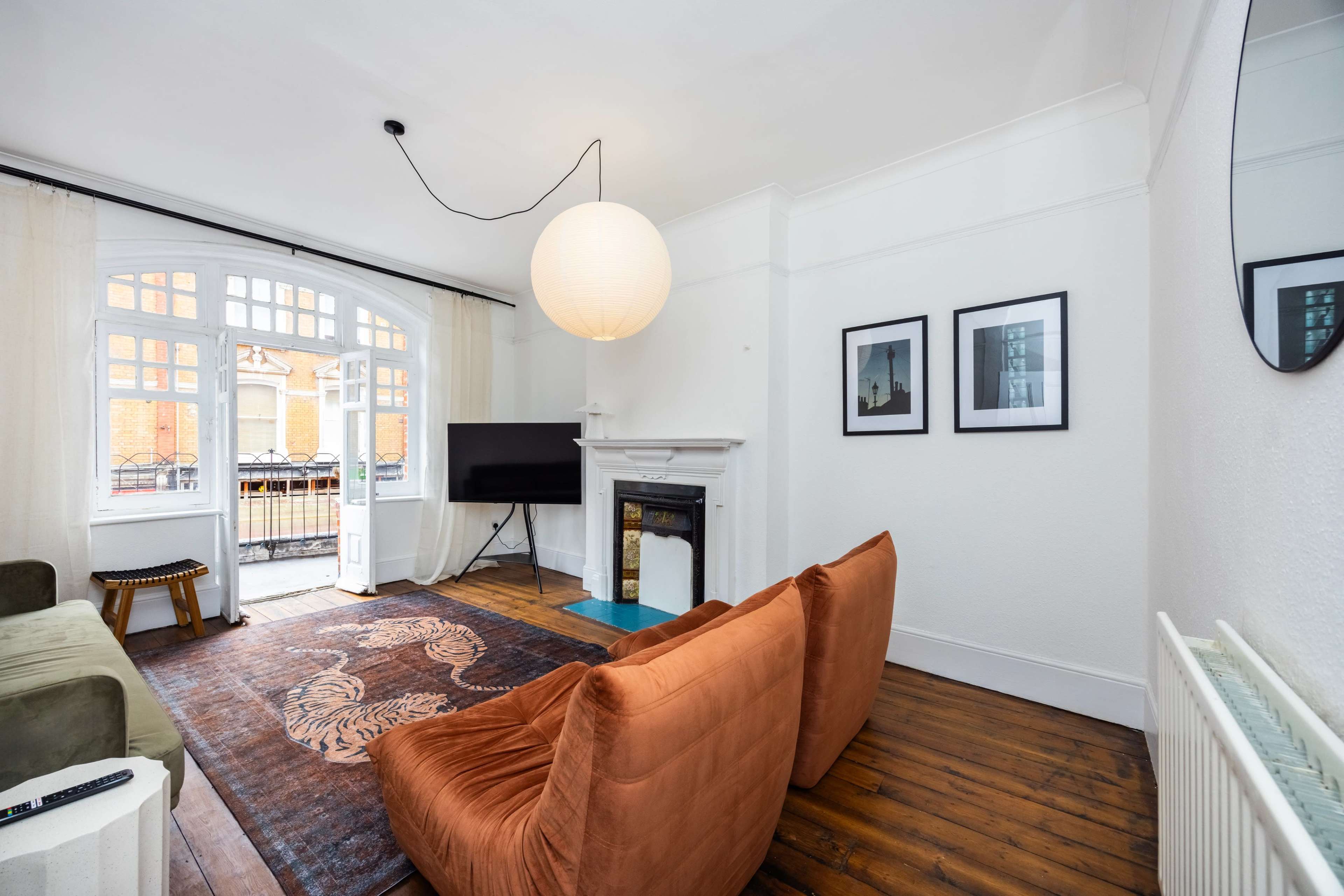 The image shows a bright living room with a pair of orange chairs, a television, and framed pictures on the walls, along with a fireplace and large windows that open to a balcony.