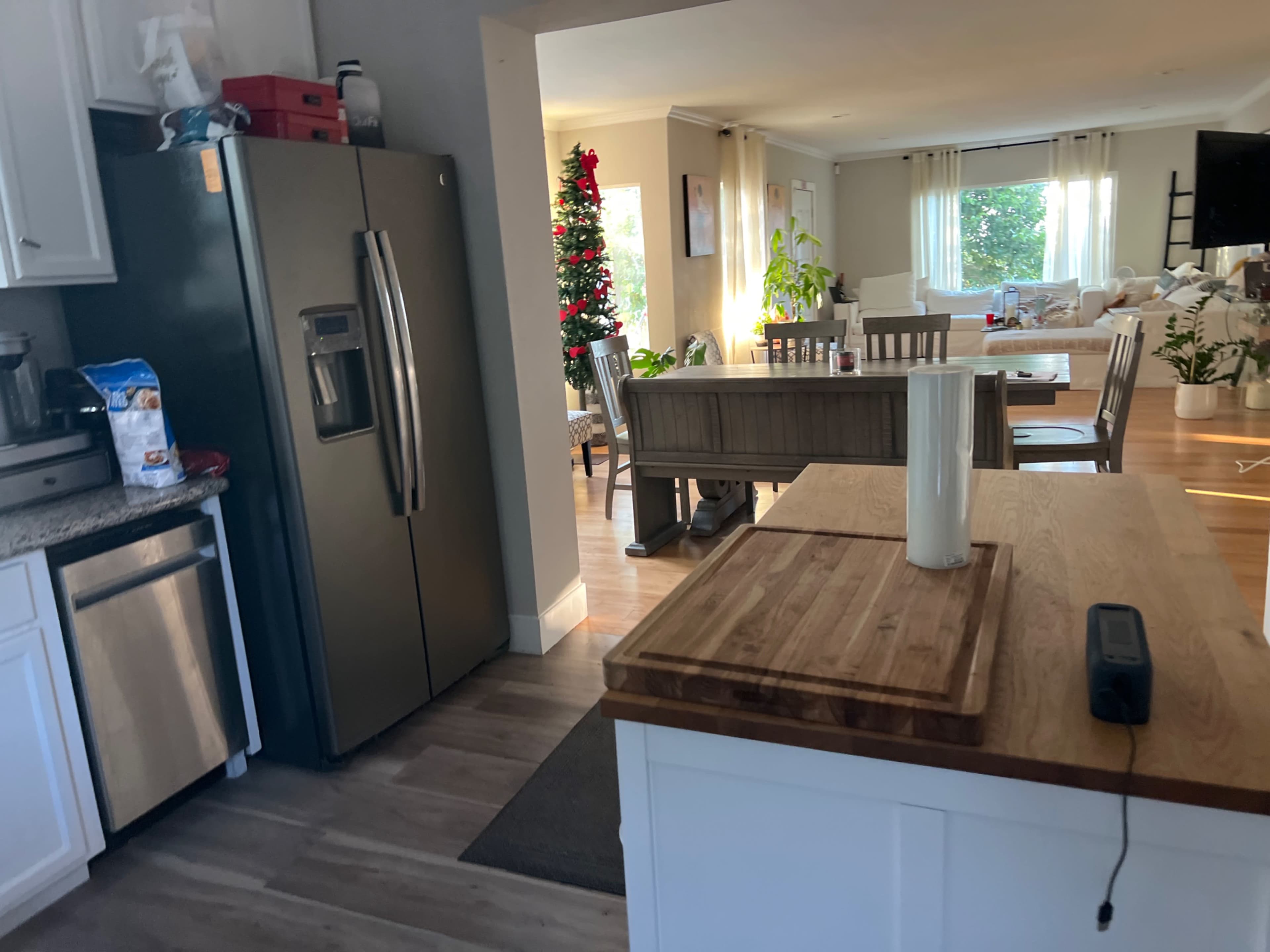 A modern kitchen features stainless steel appliances, a wooden cutting board, and connects to a dining area with a Christmas tree in the background.
