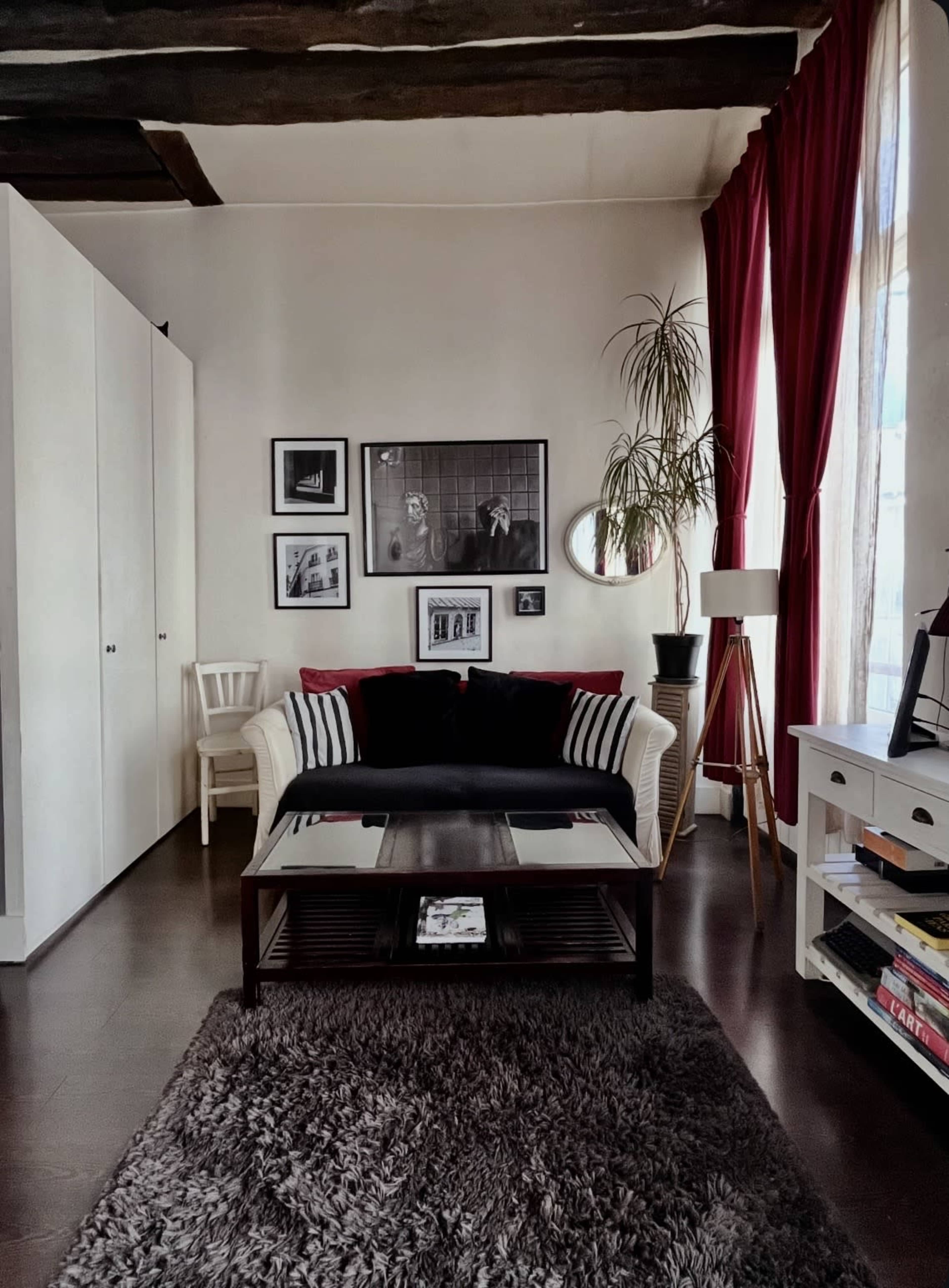 The image shows a cozy living room with a black and white couch, a coffee table, a textured rug, framed photographs on the wall, and large windows draped in red curtains.