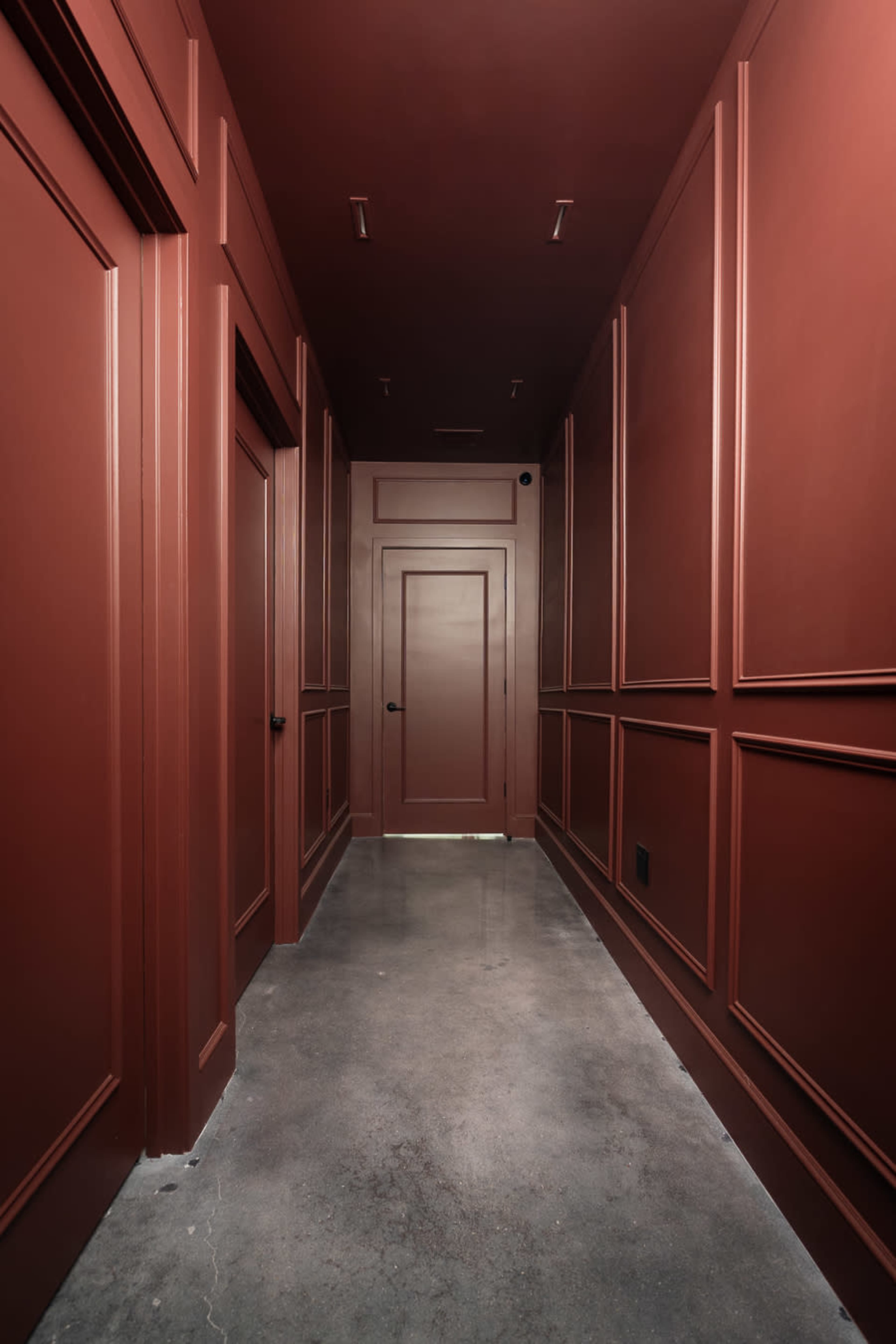 The image shows a narrow hallway with red paneled walls and concrete flooring, featuring several closed doors on either side.