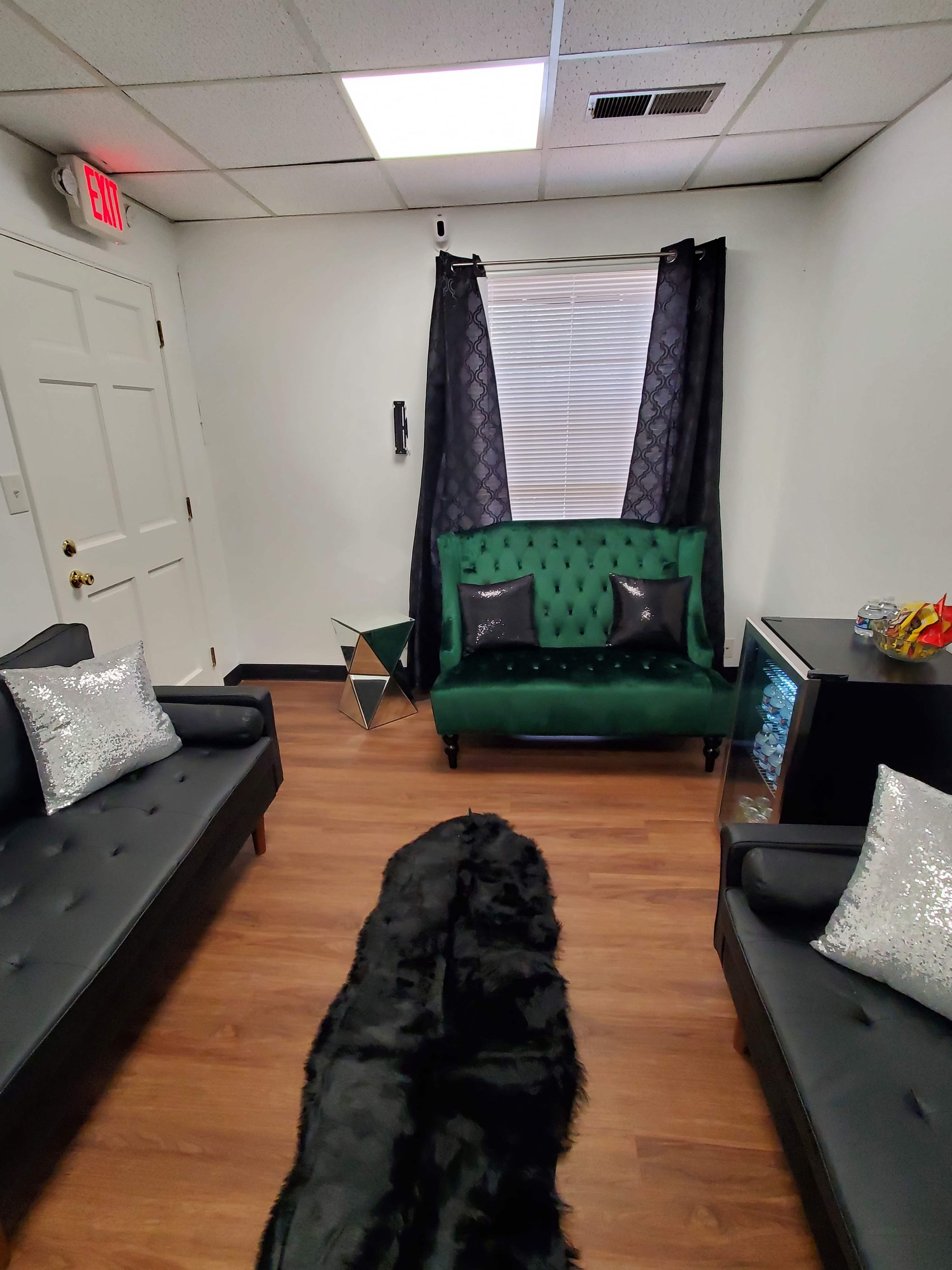 The image shows a small waiting room with a green upholstered loveseat, two black couches, a mirrored decorative object, and a black rug on the wooden floor.