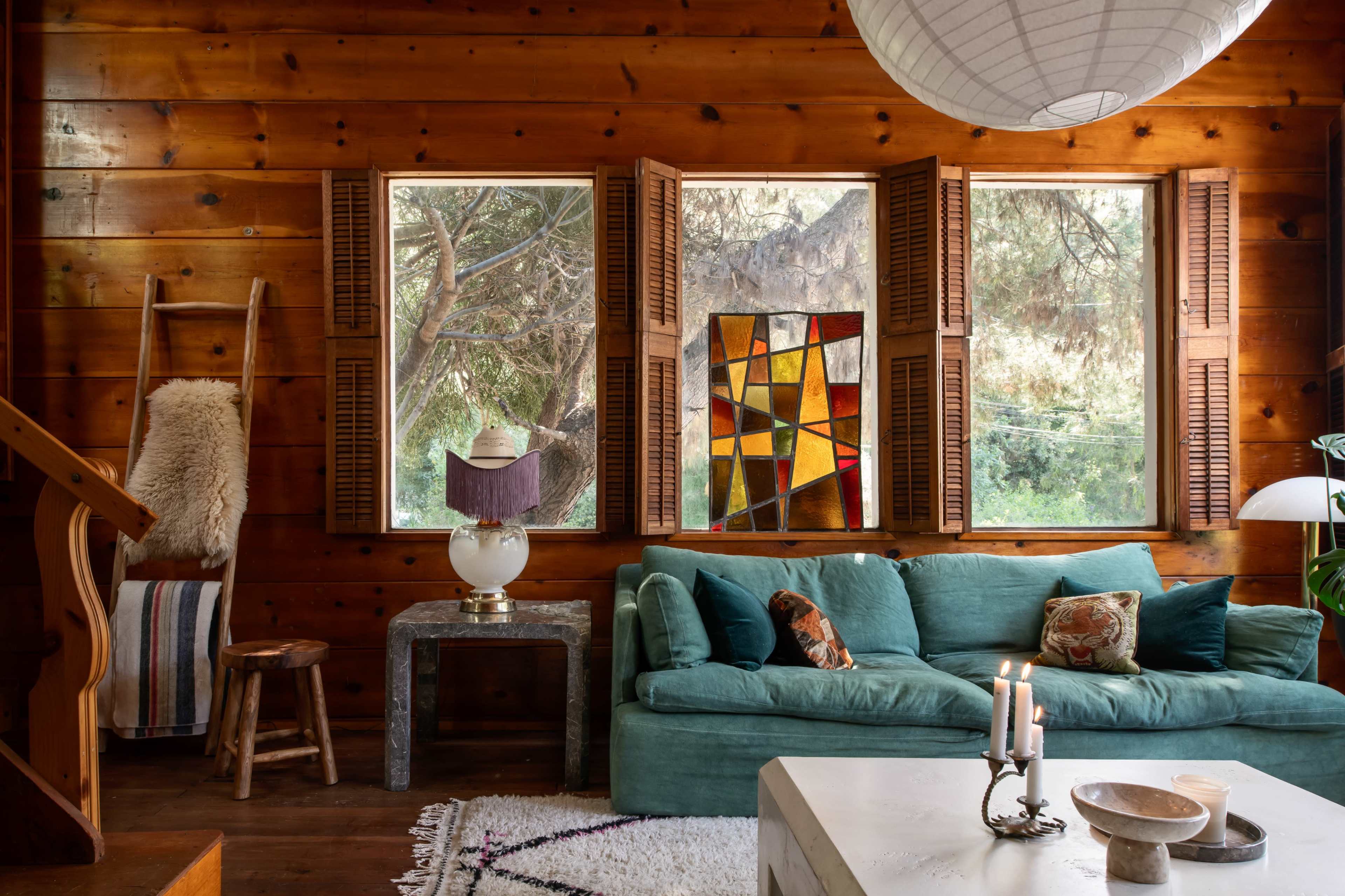 A cozy wooden living room with large windows featuring stained glass, a teal sofa, and a small table with a lamp.