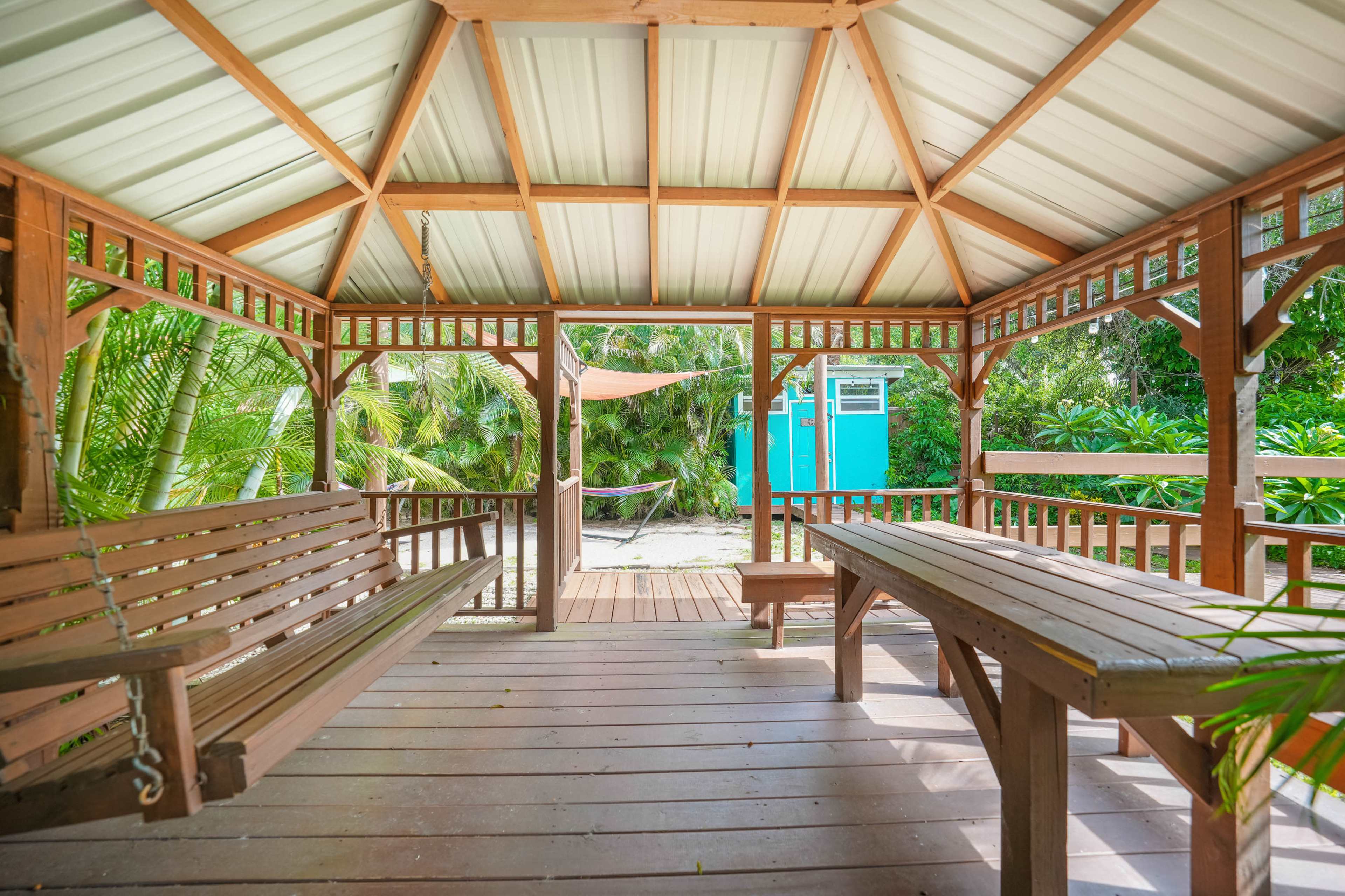 A wooden gazebo with benches and a table overlooks a garden area featuring a hammock and a blue shed.