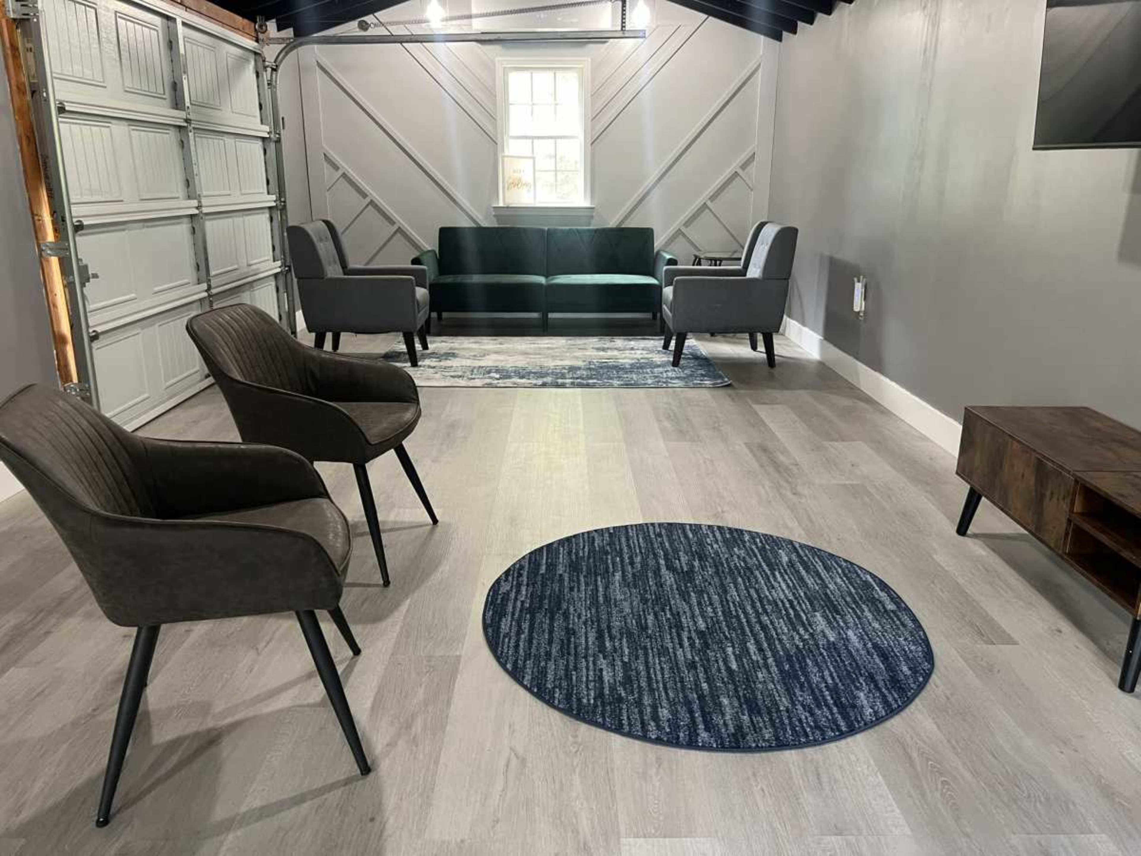 The image shows a modern interior space featuring two armchairs and a sofa arranged around a circular rug, with a wooden TV stand against one wall.
