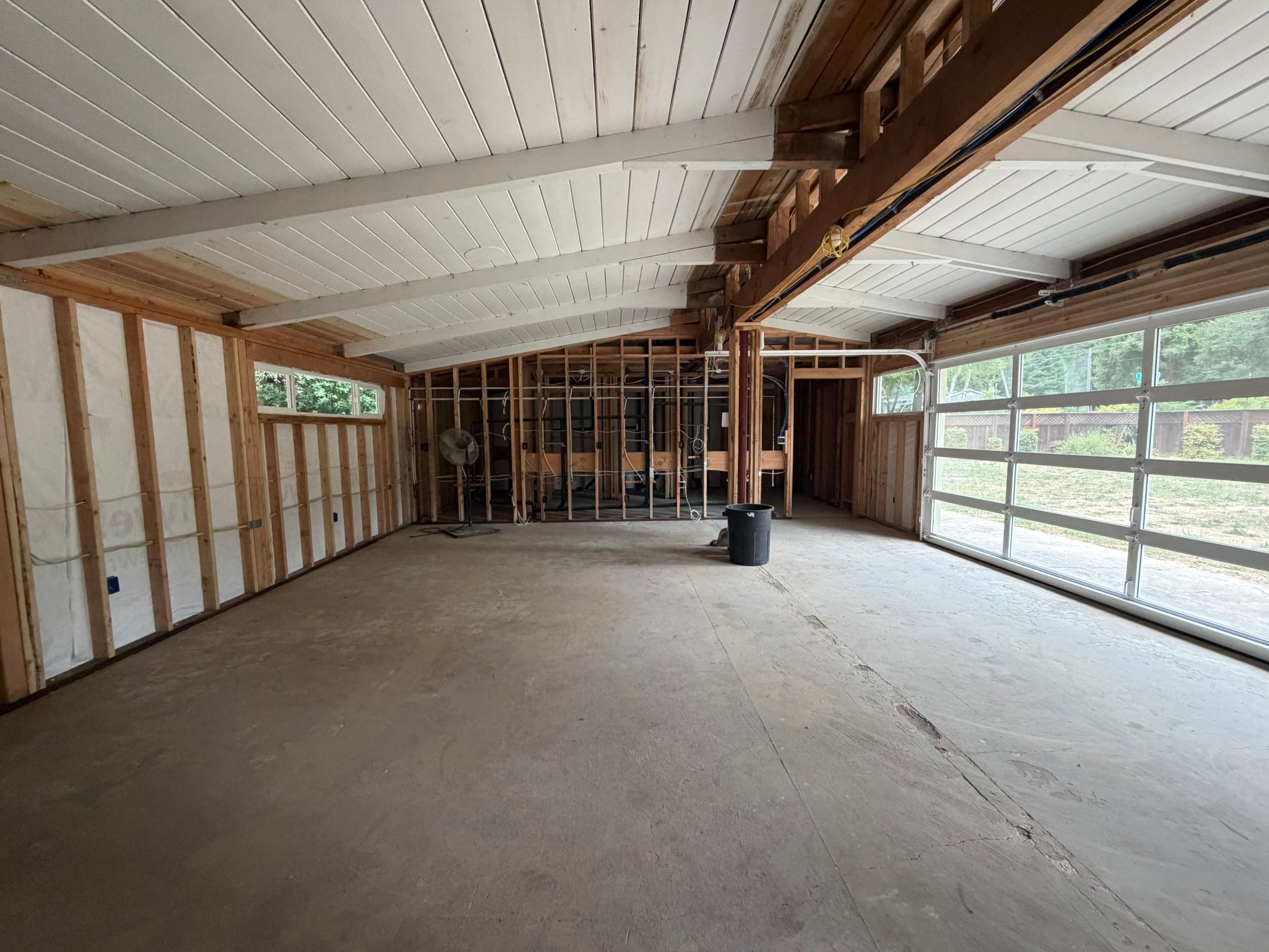 The image shows an empty room with exposed framing, a concrete floor, and large windows along one wall.