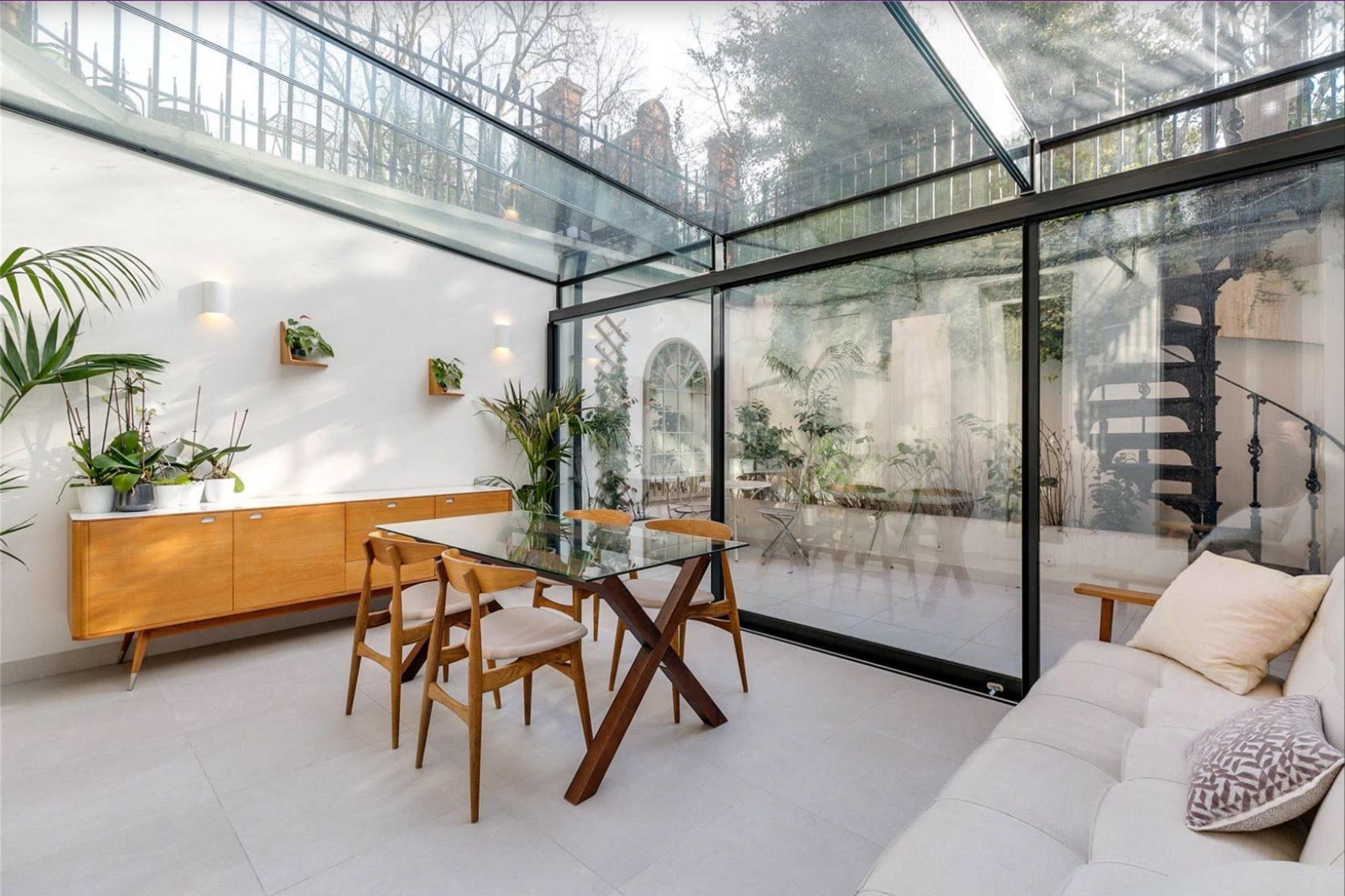 A bright, modern sunroom with a glass ceiling, featuring a wooden dining table and chairs, a sideboard, and various indoor plants.