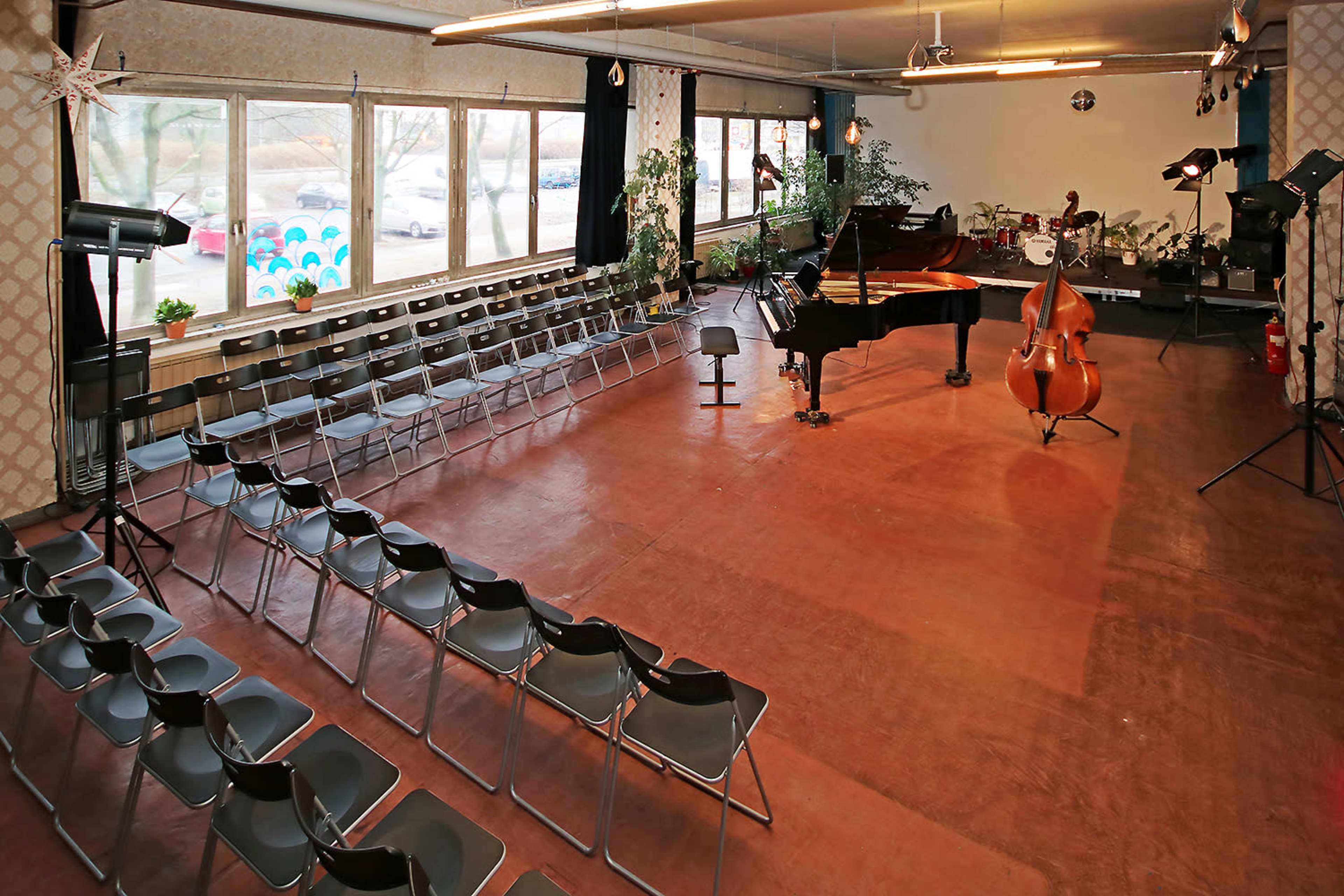 A music performance space features rows of black chairs facing a grand piano and double bass, with large windows providing natural light.
