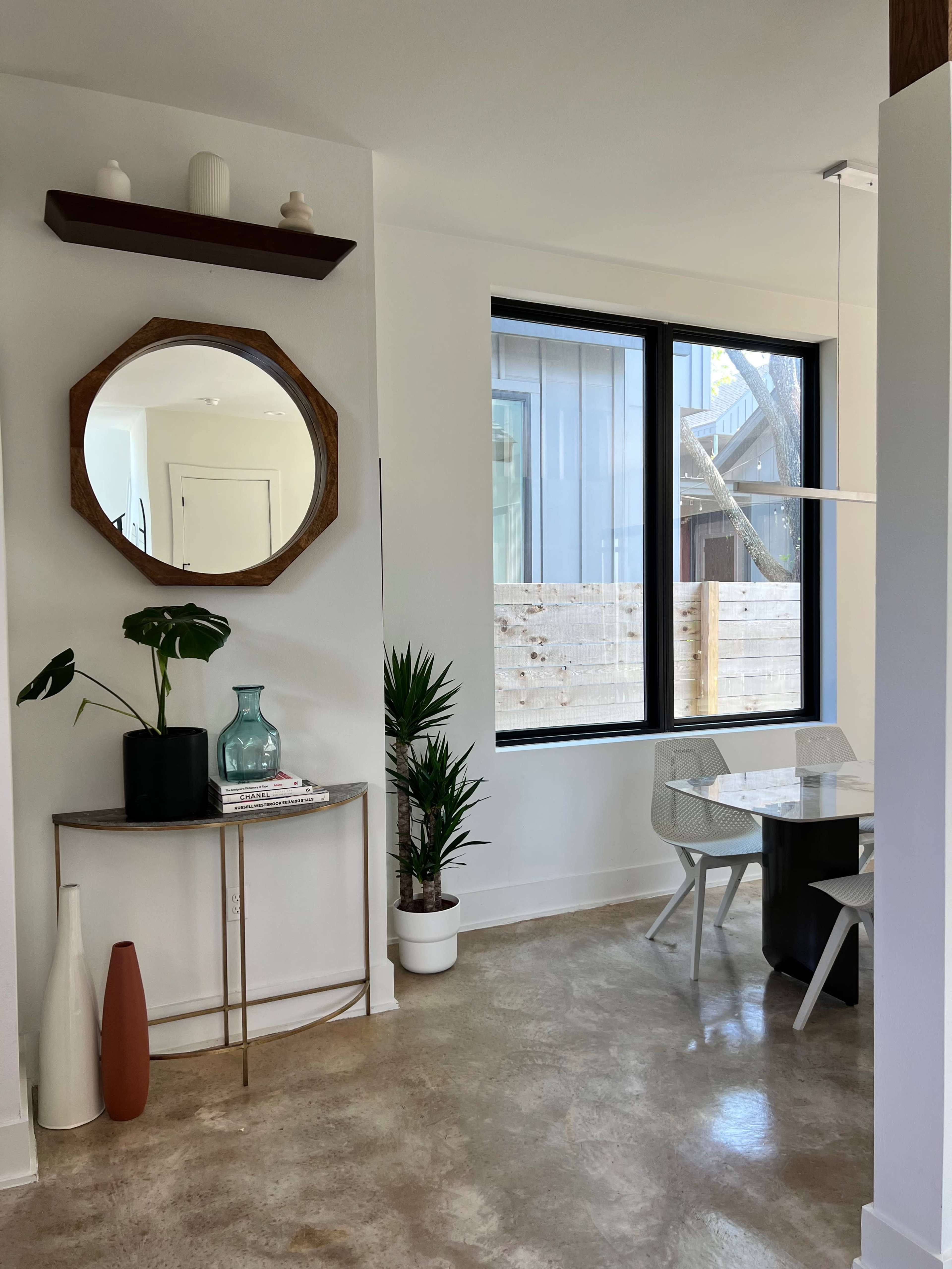 A modern dining area with a round table, a set of white chairs, a decorative console table with plants, and large windows allowing natural light.