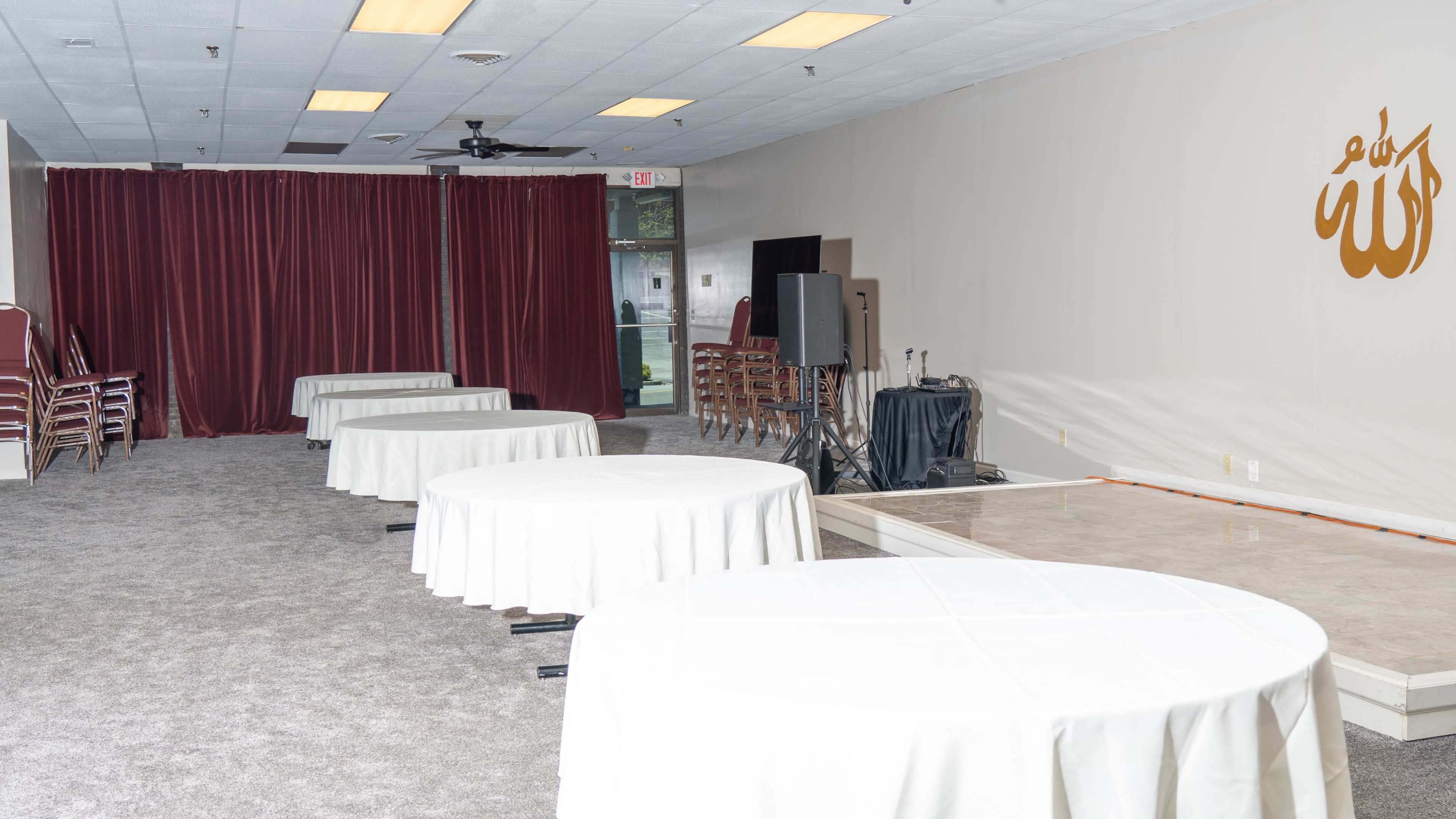 A room set up with several round tables covered in white tablecloths, a stage area, and red curtains in the background.