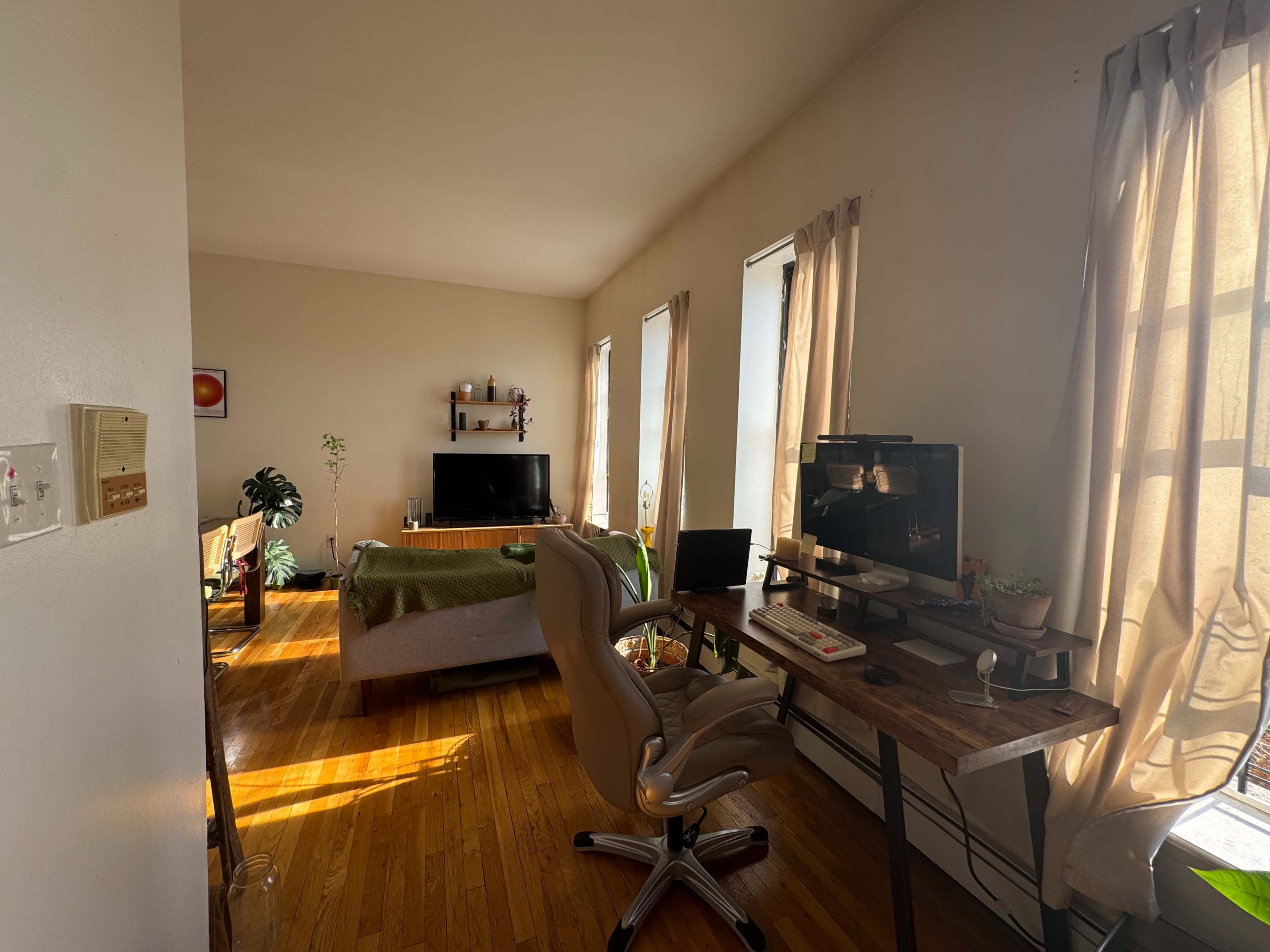 The image shows a bright living room with a TV, a green couch, a desk with a chair, and large windows draped with curtains.