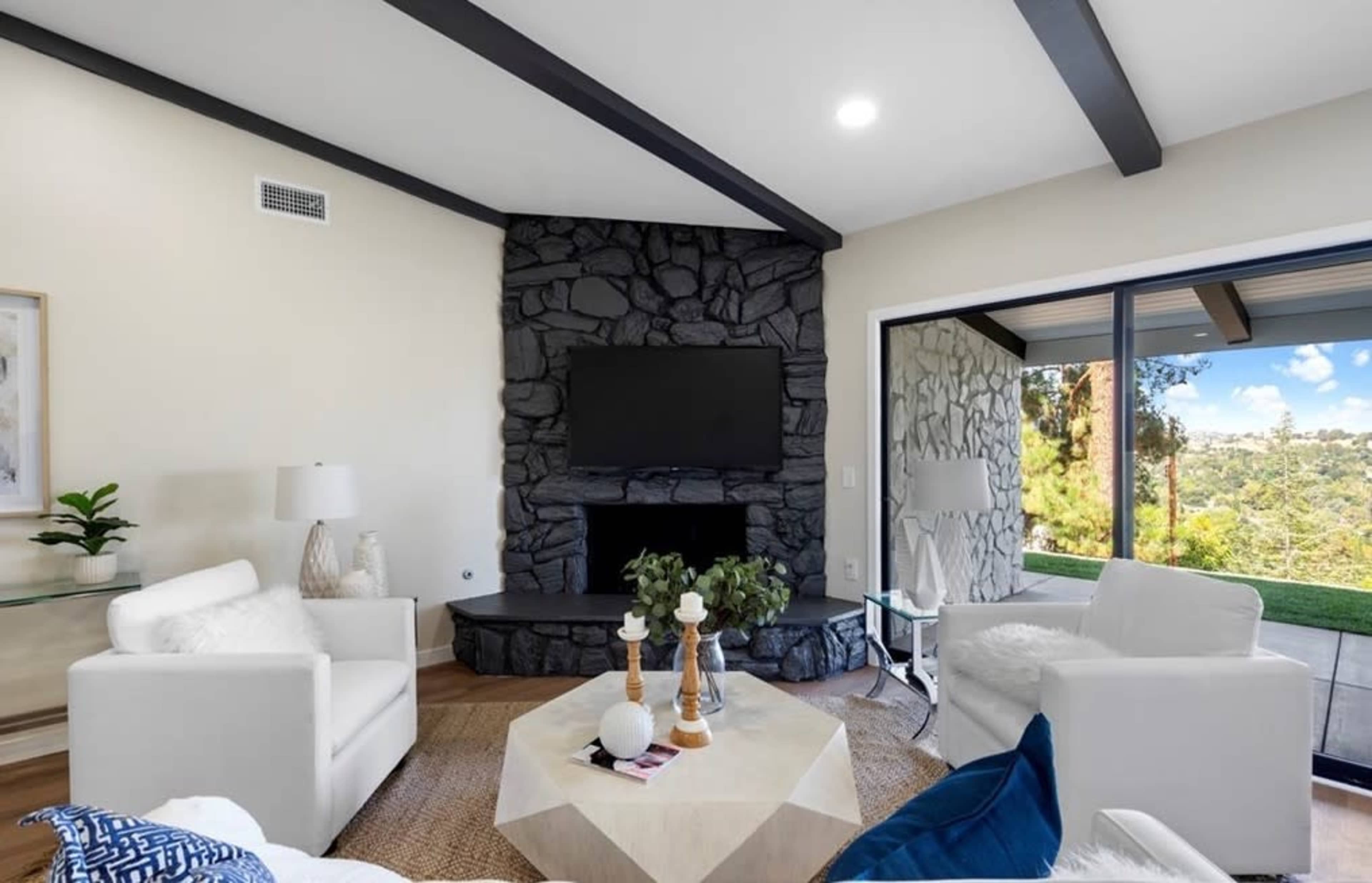 A modern living room features a stone fireplace, a flat-screen TV, and large windows overlooking a scenic view.