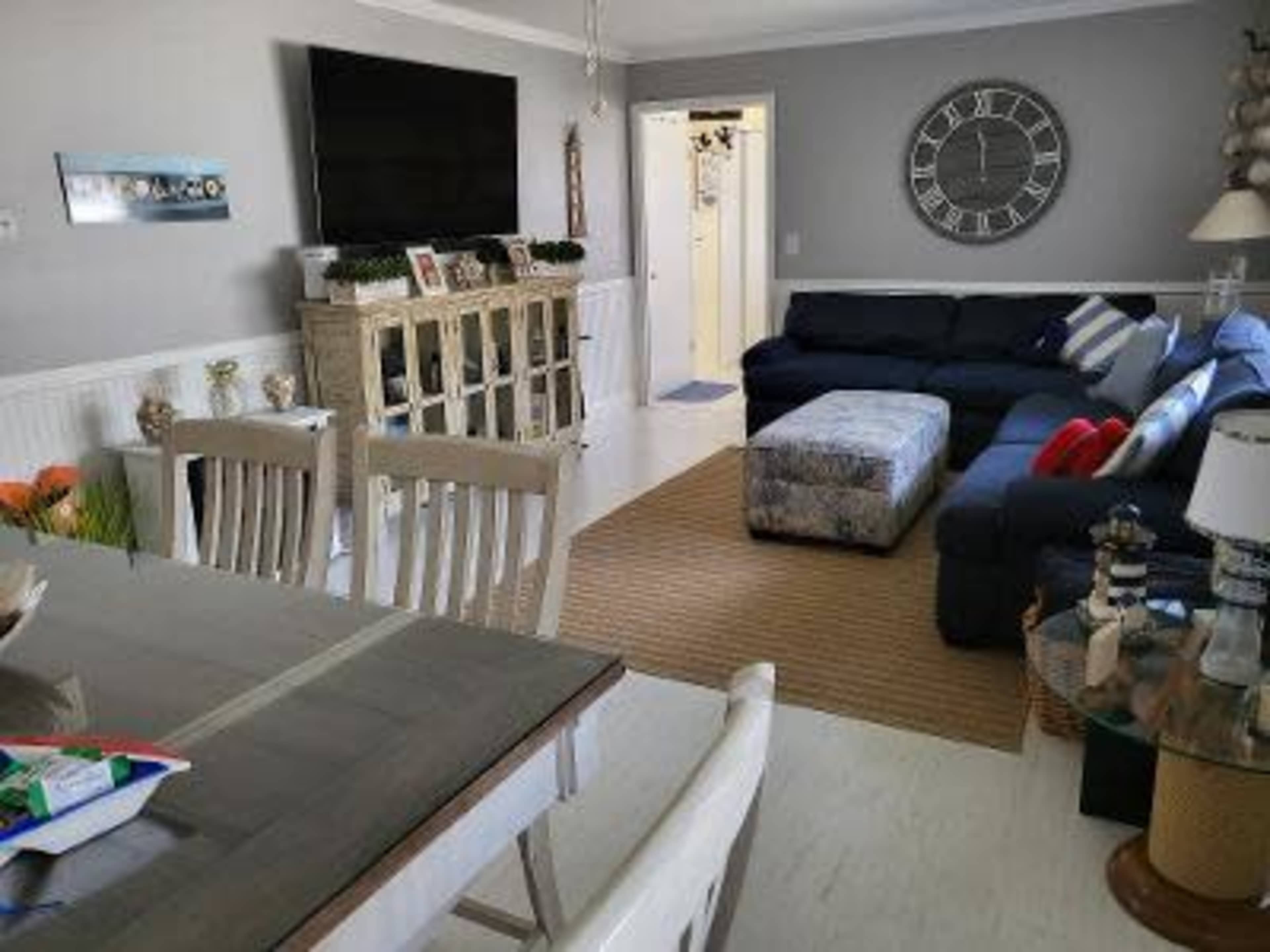 A living room with a large television, a sectional sofa, an ottoman, a dining table, and a clock on the wall.