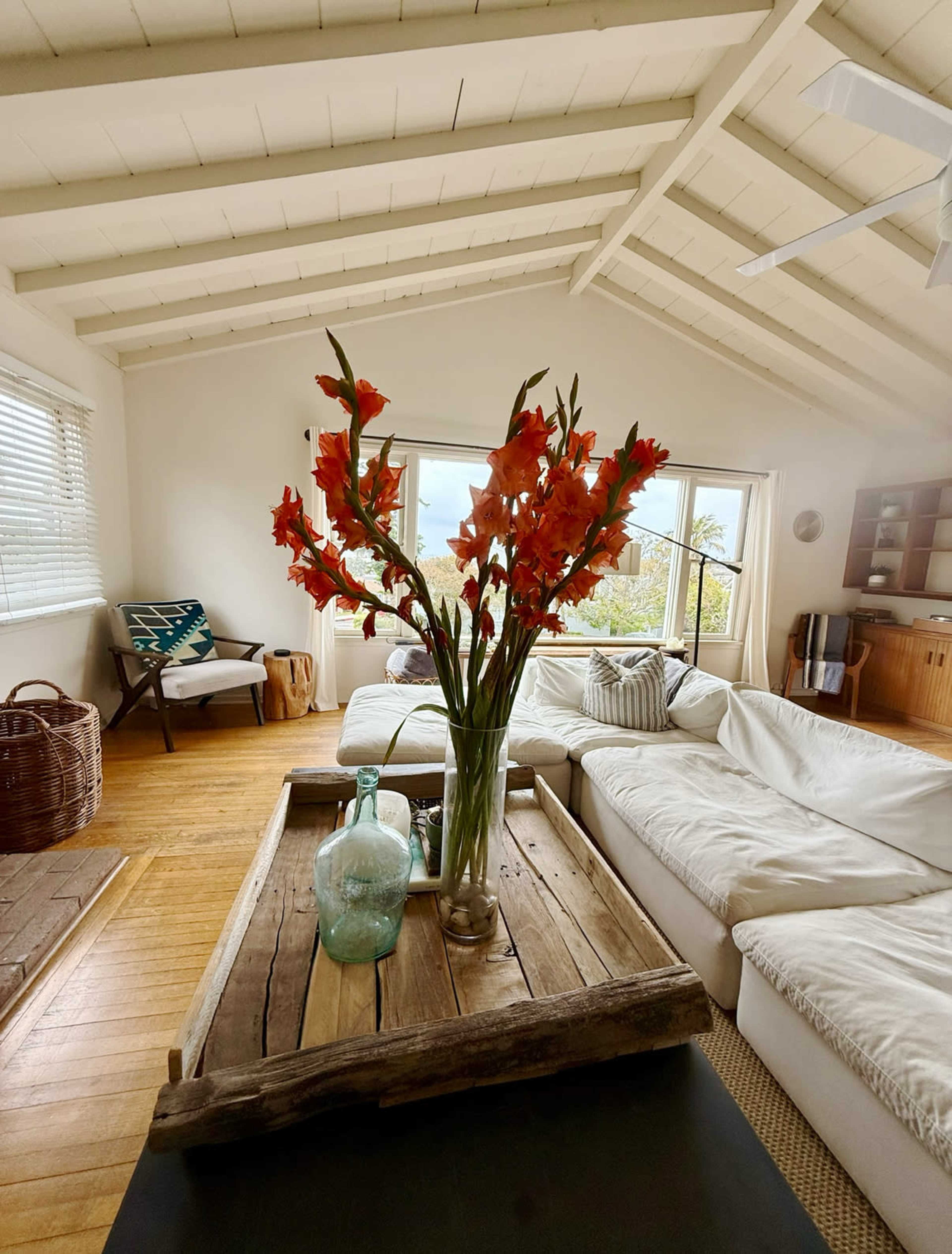 A bouquet of orange gladiolus is placed in a vase on a rustic wooden coffee table, surrounded by a bright, airy living room featuring white sofas and natural light.