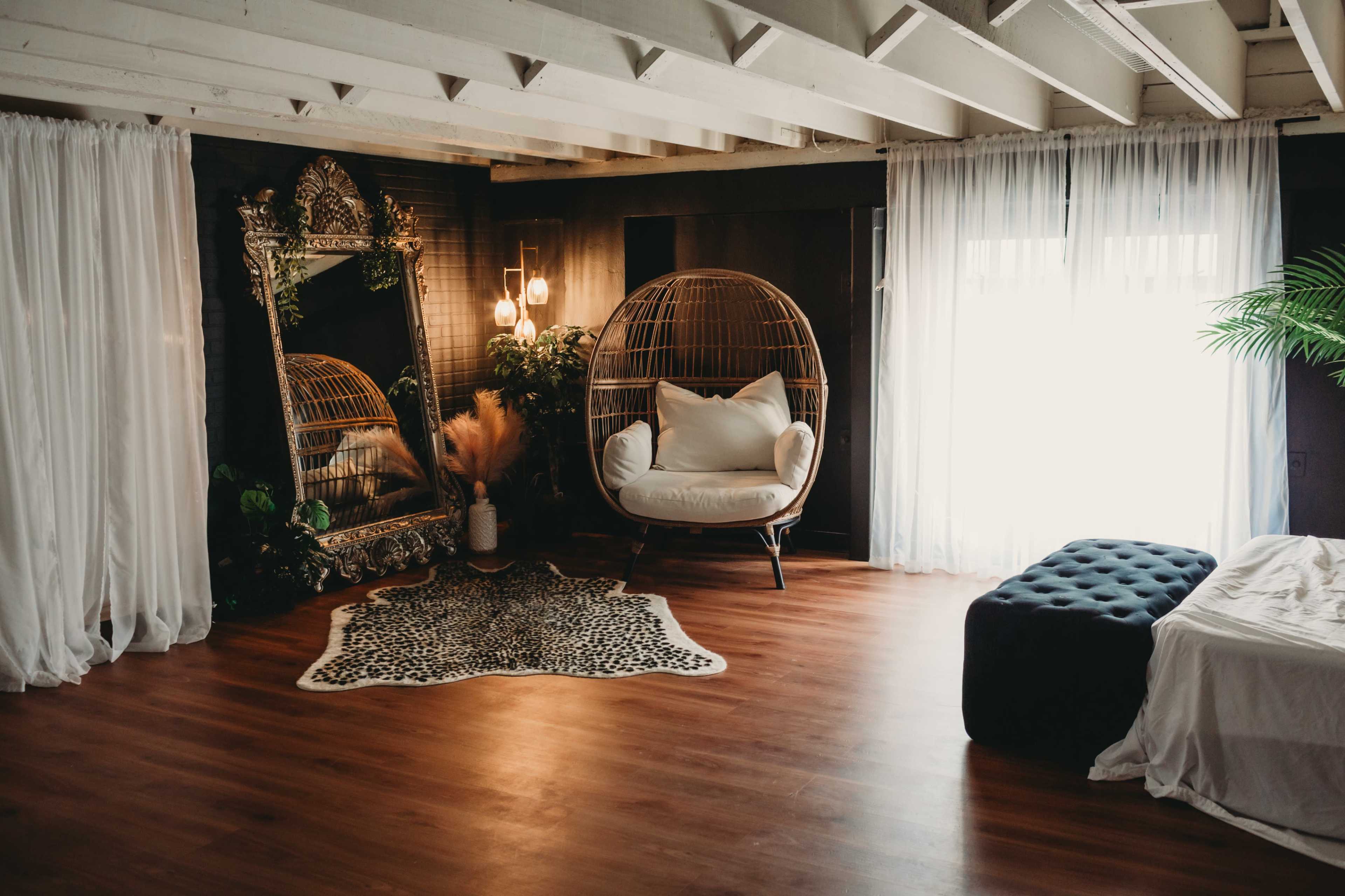 A spacious indoor area with wooden flooring, featuring a large decorative mirror, a hanging chair, and a plush ottoman, all surrounded by sheer white curtains and a decorative rug.