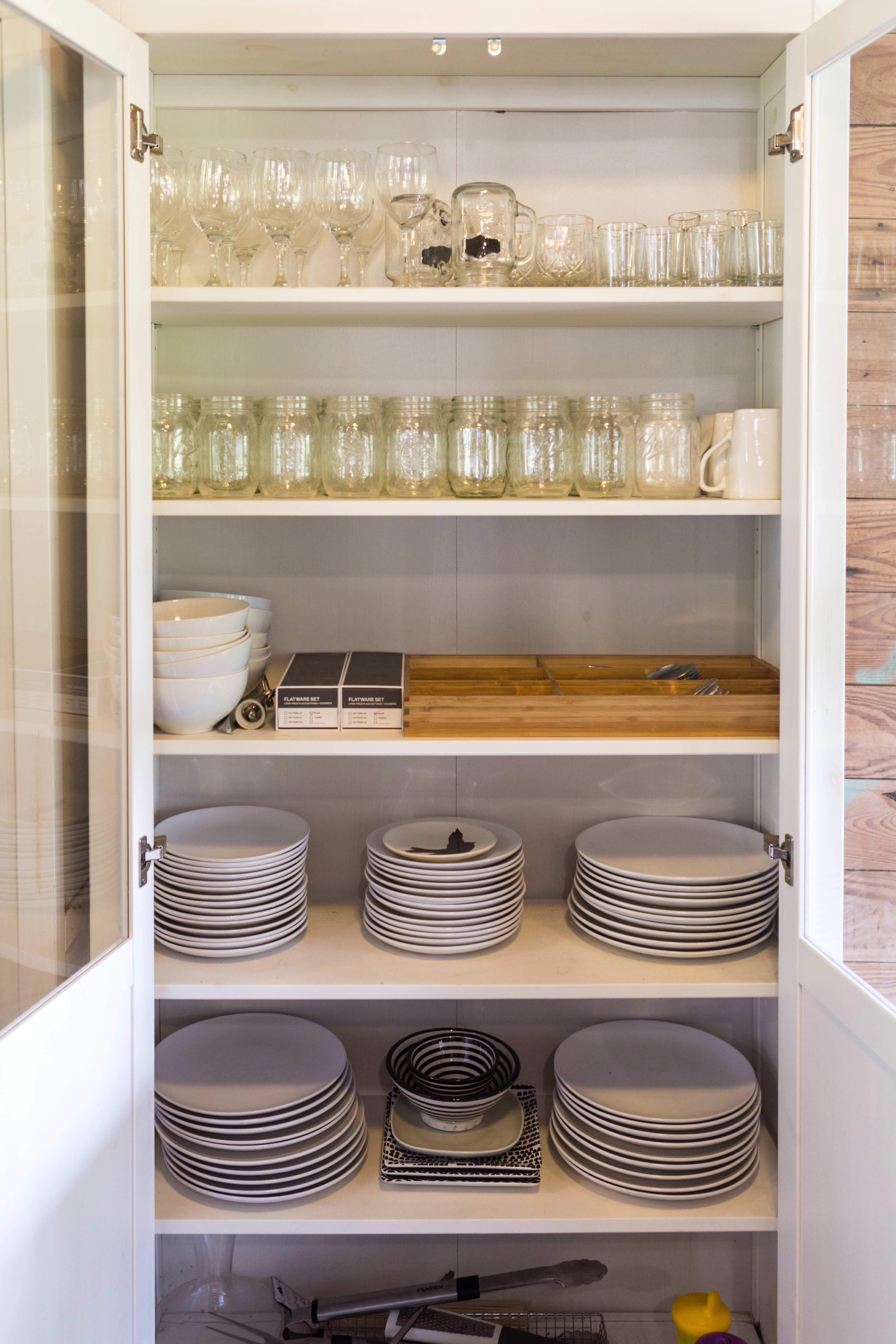 A kitchen cabinet contains neatly arranged glassware, dishes, and utensils across its shelves.