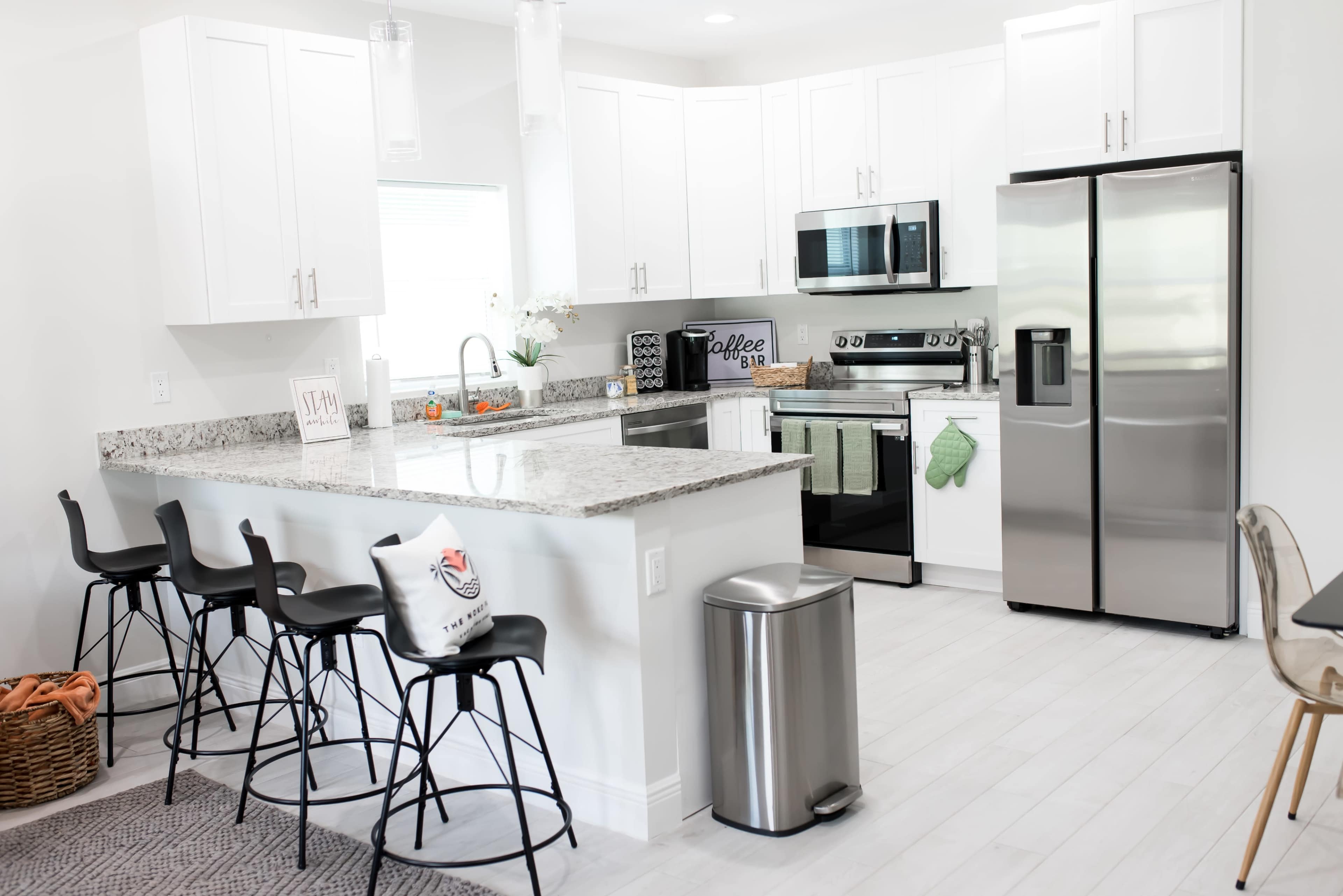 A modern kitchen features white cabinets, stainless steel appliances, and a granite countertop with bar stools at a breakfast bar.