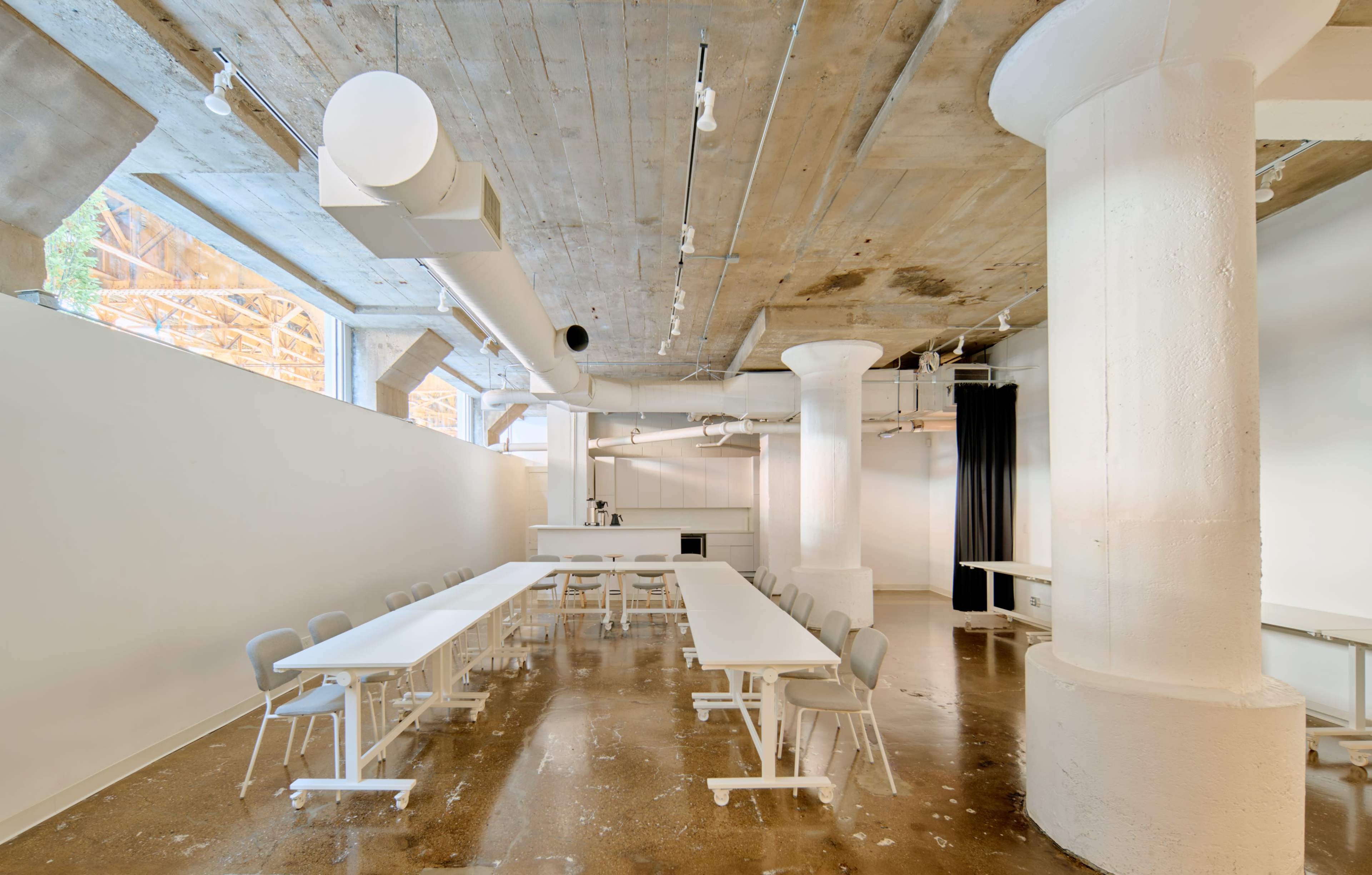 The image shows a spacious room with polished concrete floors, large white pillars, and a long table set up for meetings or collaboration.