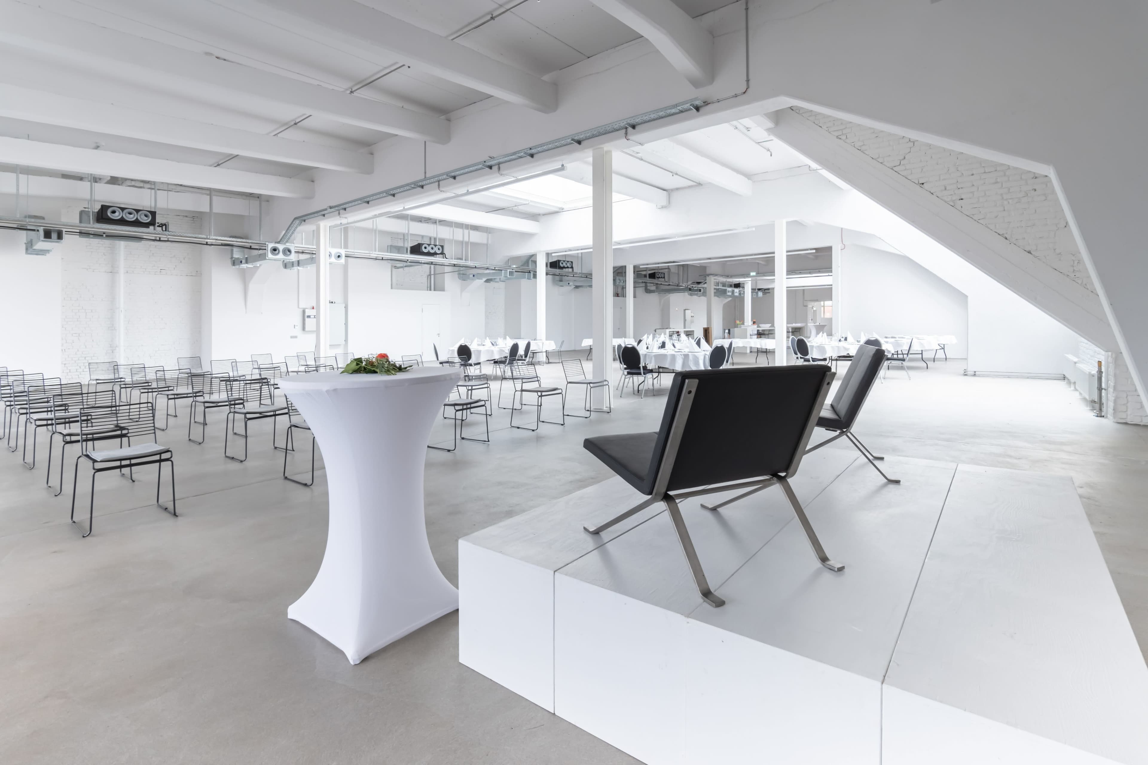A spacious, modern event room features rows of black chairs, a small raised platform with two black chairs, and several round dining tables set for an event.