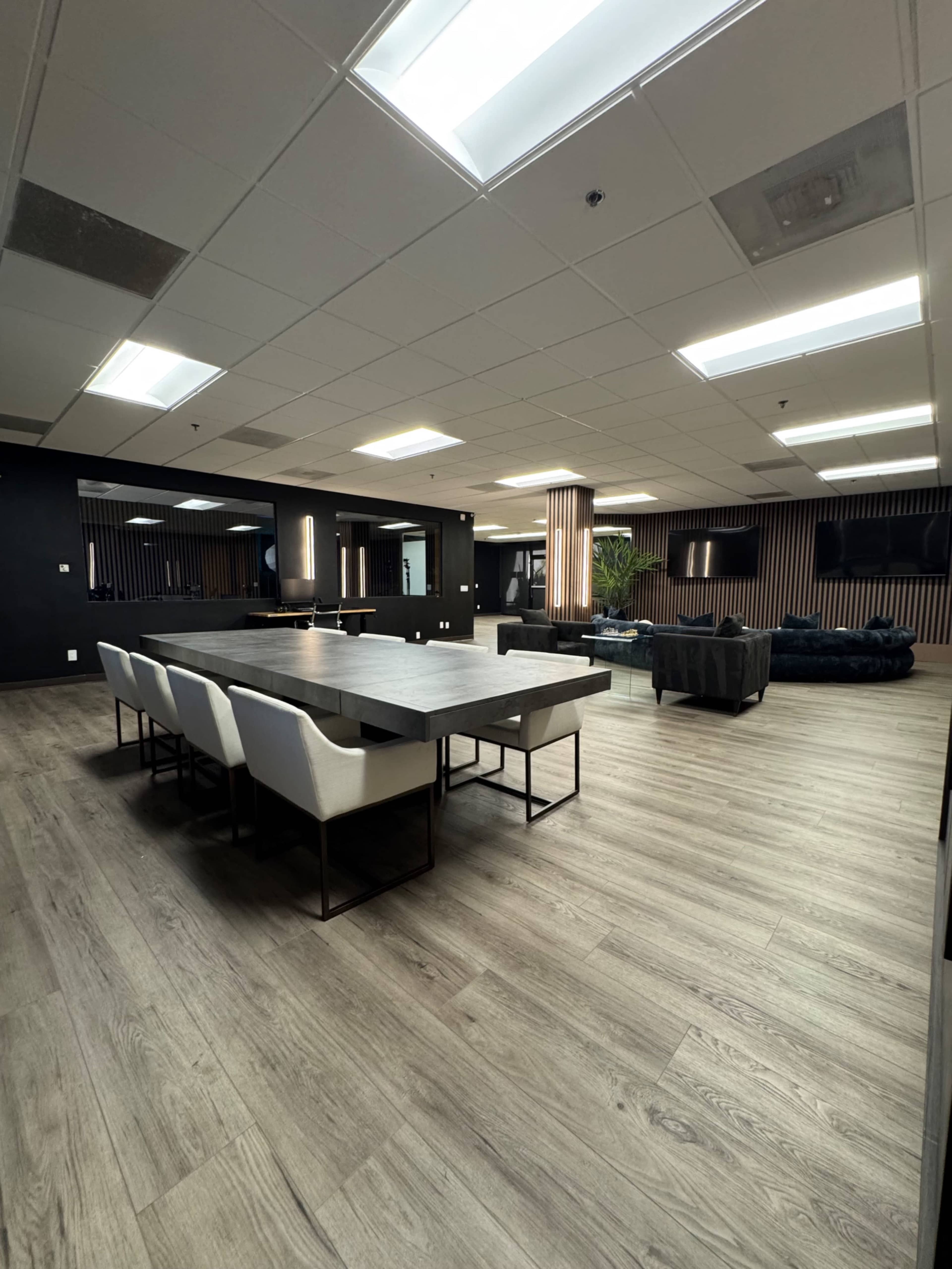 The image shows a modern office space with a large wooden table surrounded by white chairs, dark walls, and a lounge area with a couch in the background.