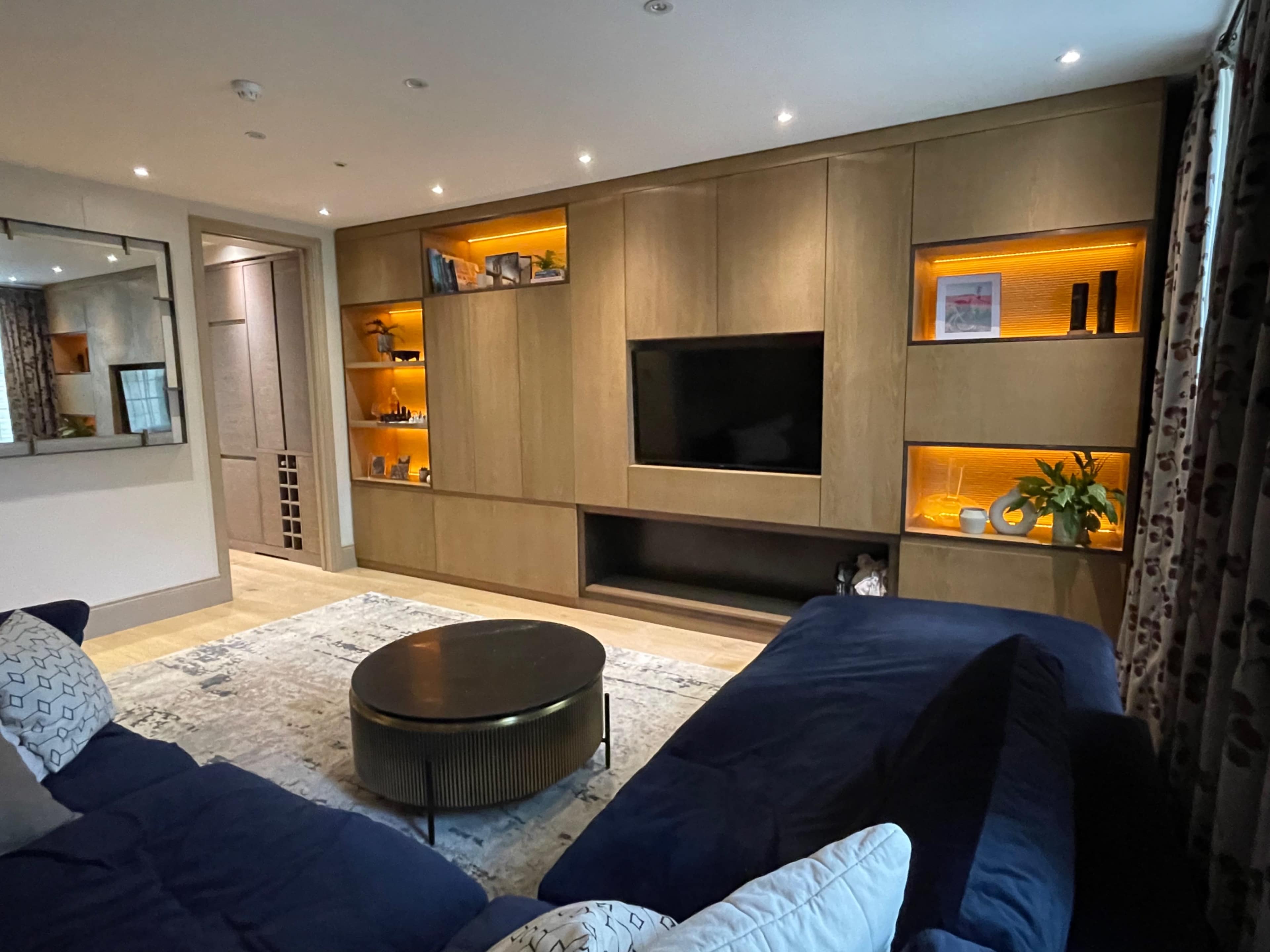The image shows a modern living room featuring a dark blue sectional sofa, a round coffee table, and a wooden entertainment unit with backlit shelves.