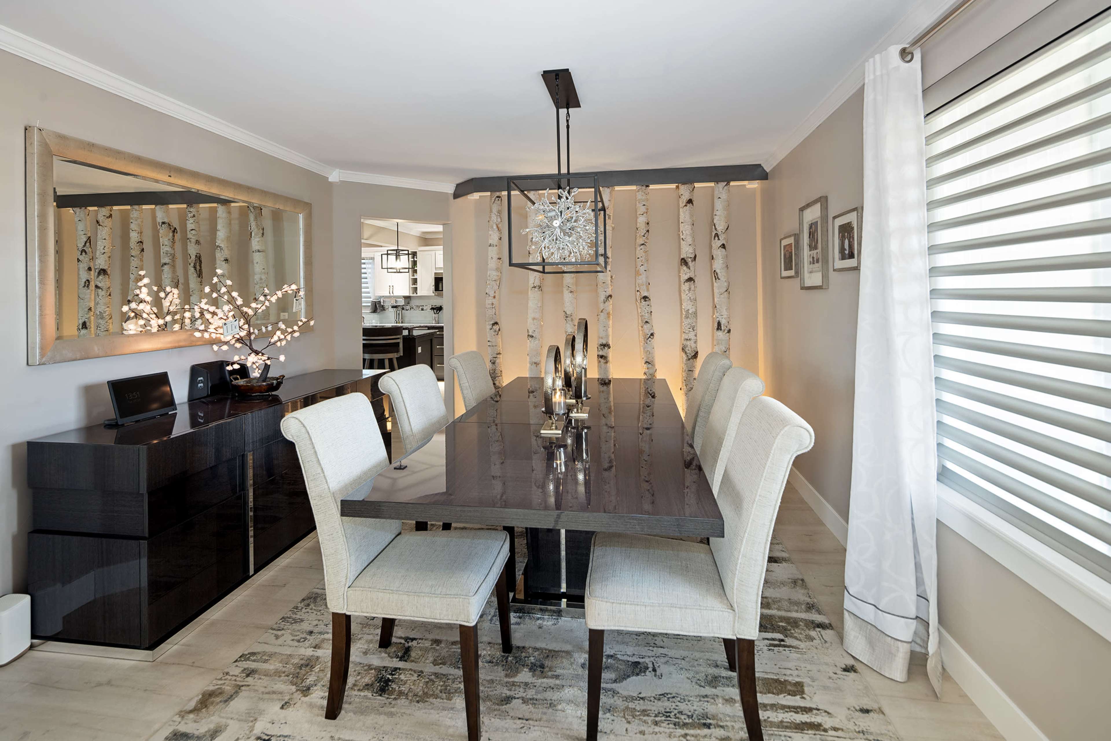 The image shows a modern dining room featuring a long wooden table surrounded by upholstered chairs, with an elegant light fixture above and a mirrored wall in the background.