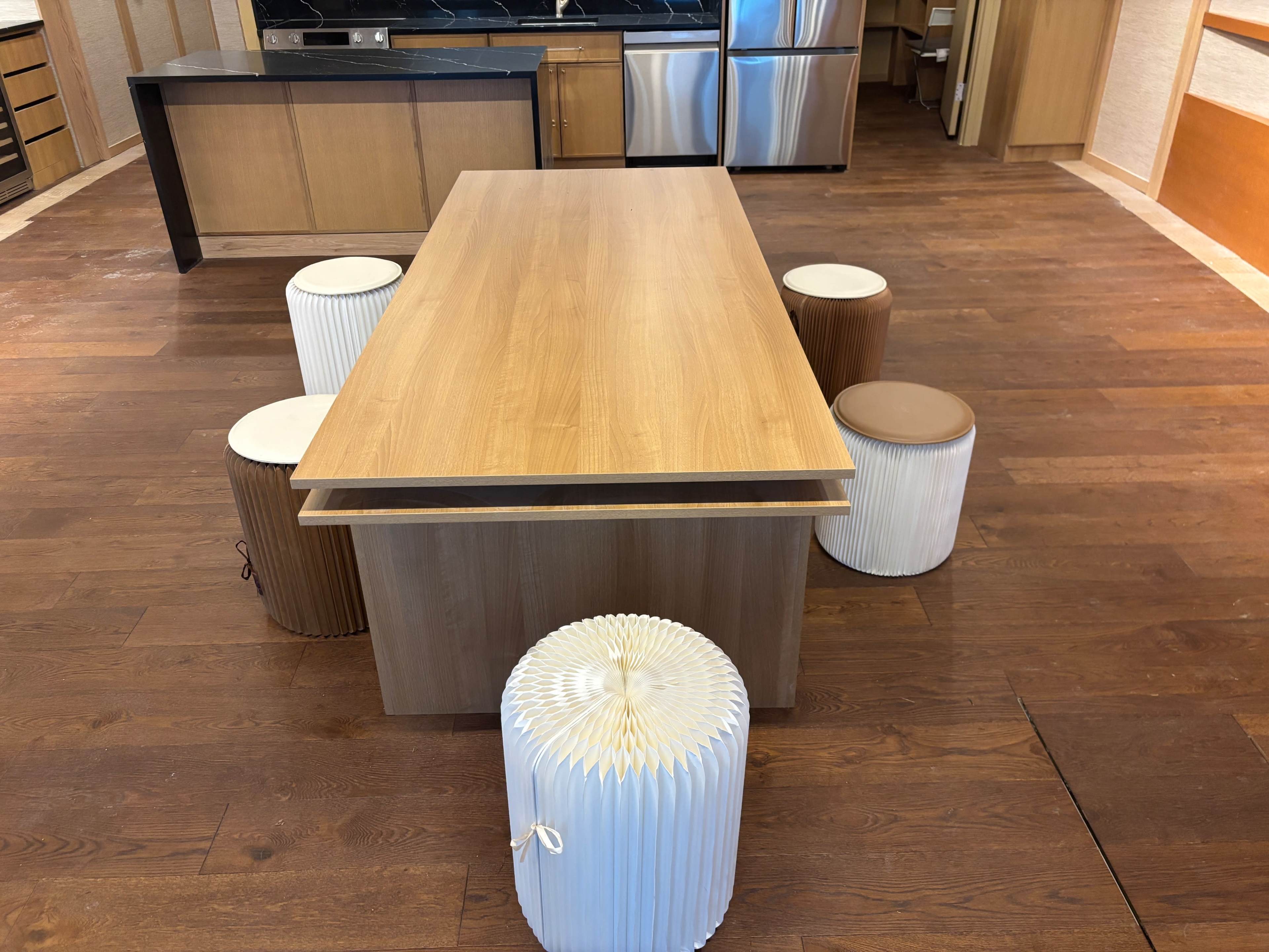 A wooden table is placed in the center of a kitchen area, surrounded by four cylindrical stools.