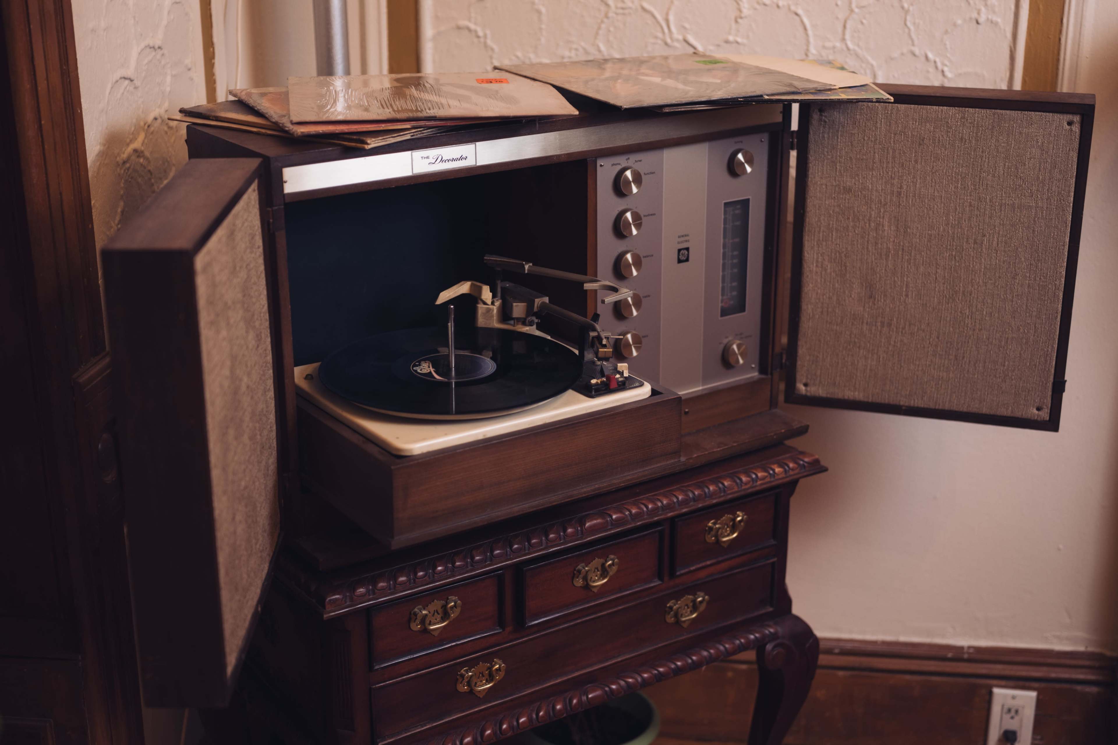 A vintage turntable with wooden cabinets is set on a carved wooden table, and several vinyl records are resting on top of it.