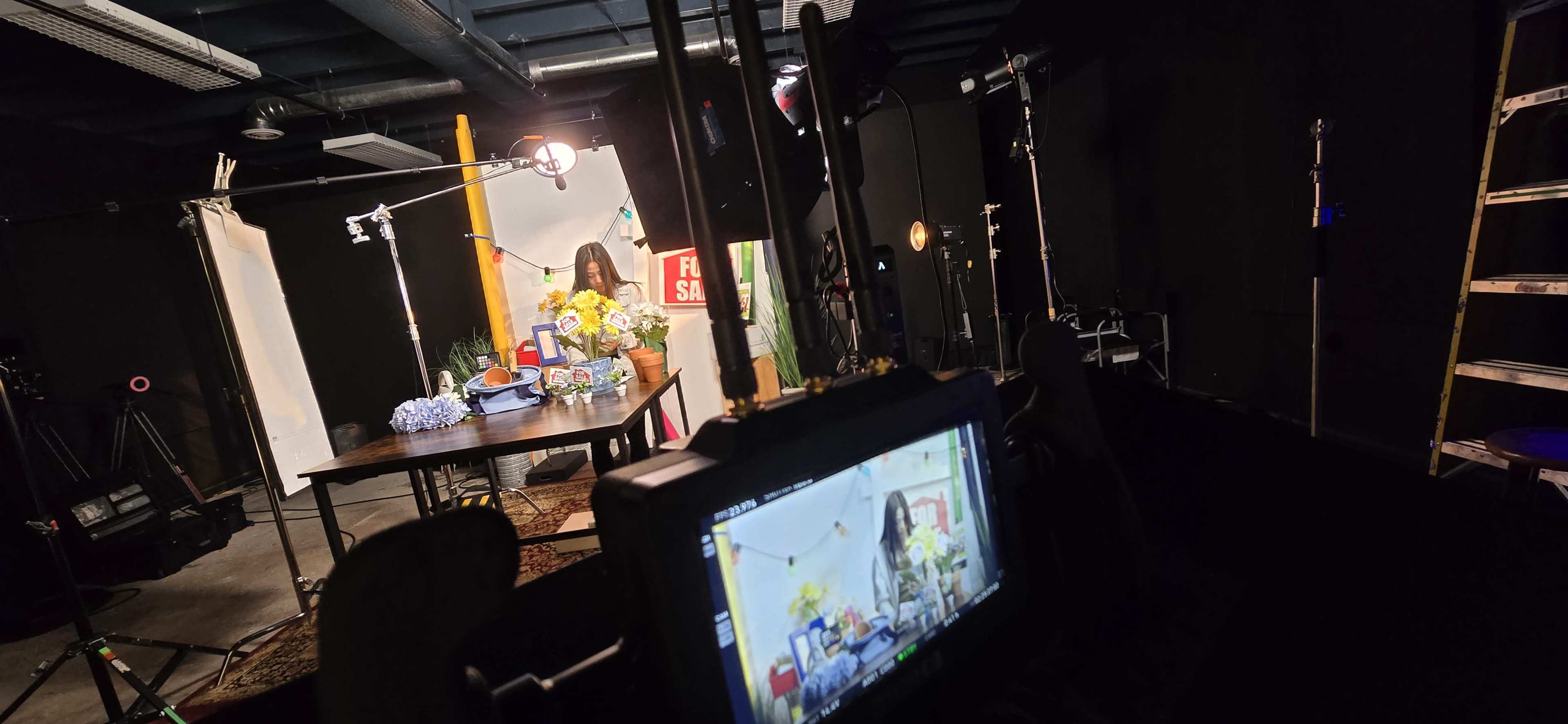 The image shows a film set with a table adorned with flowers and props, while a camera monitor displays a live feed of the scene being filmed.