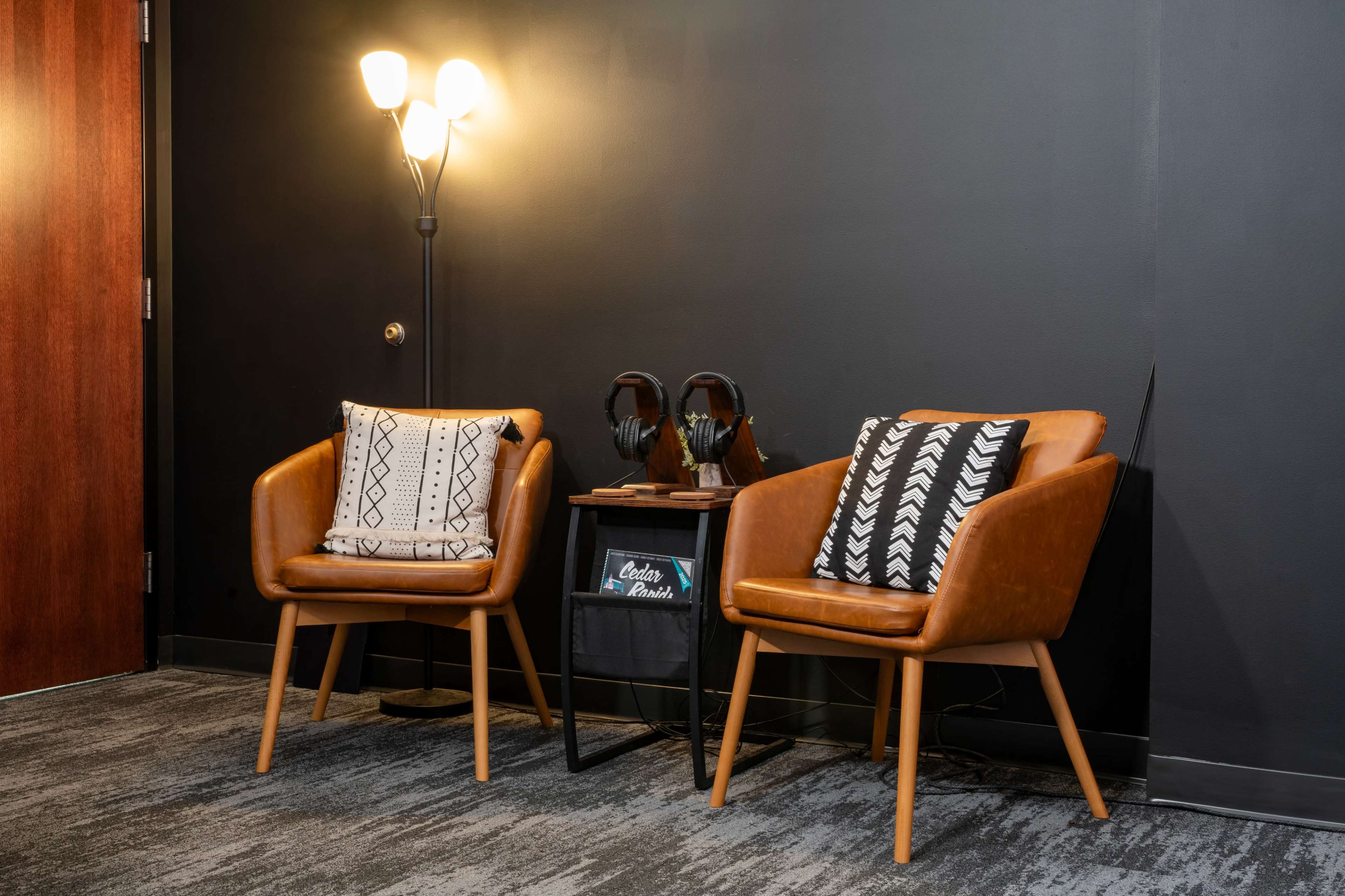 Two brown chairs with patterned cushions are placed beside a small side table, which holds a pair of headphones, next to a wall illuminated by a floor lamp.