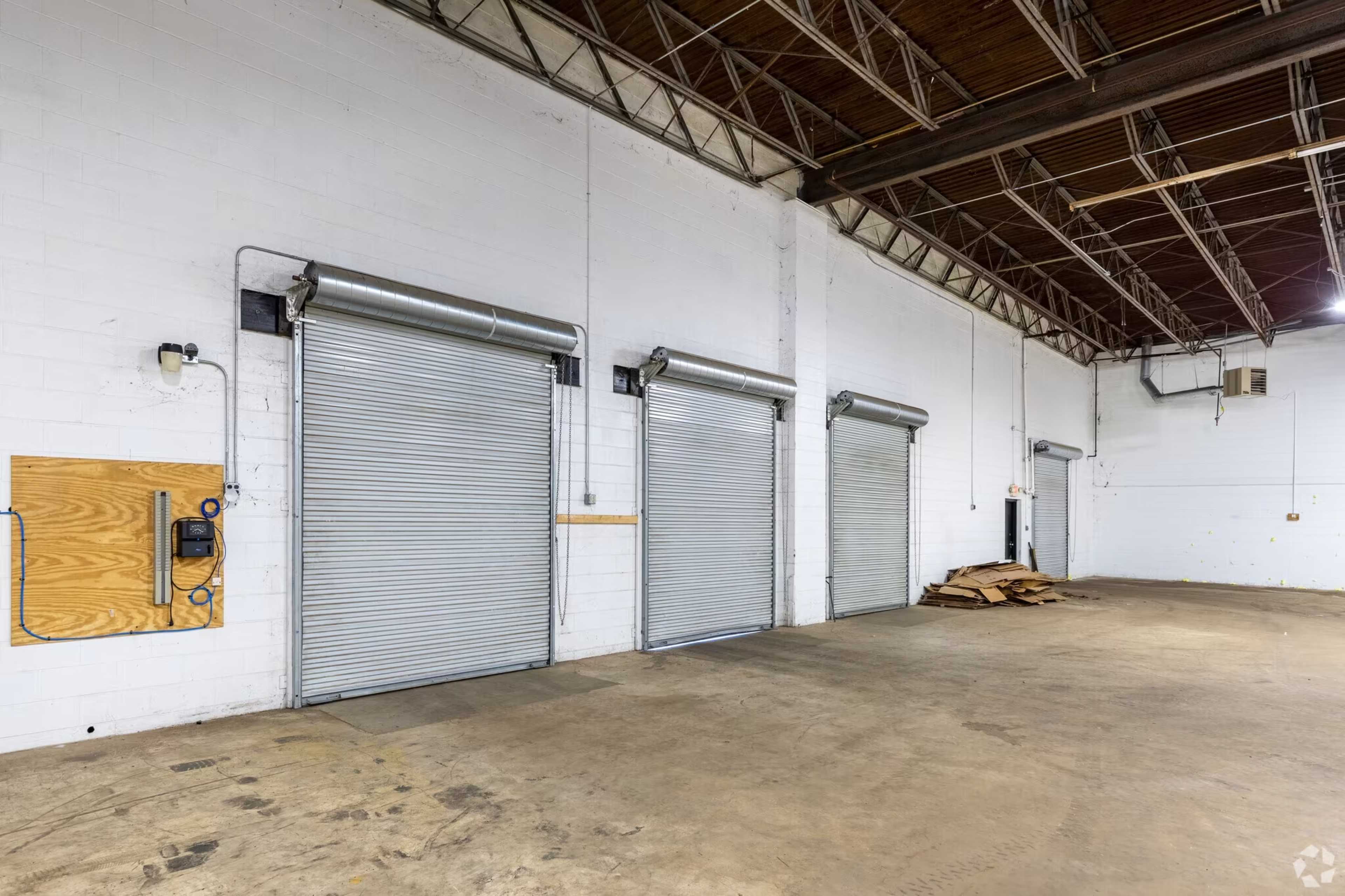 An empty warehouse space with three metal roll-up doors on a white brick wall and a concrete floor.