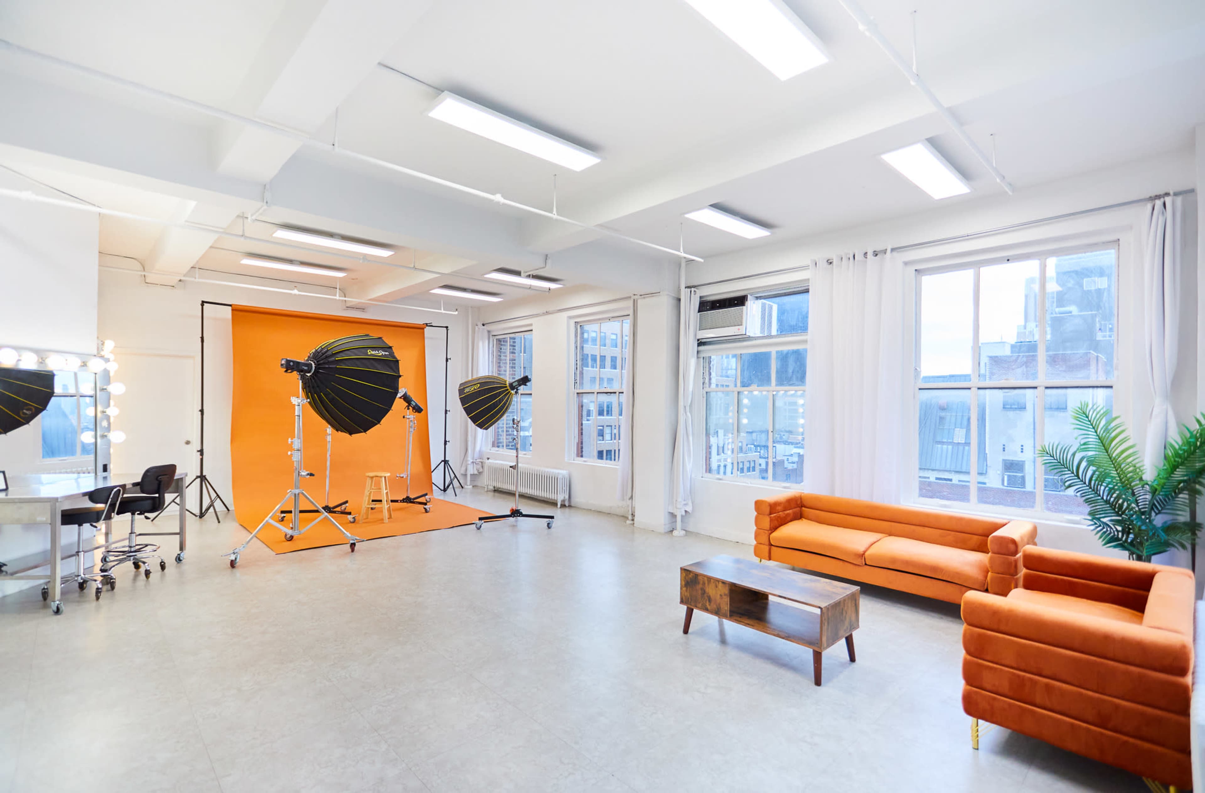 The image shows a bright photography studio with an orange backdrop, softbox lighting equipment, a seating area with an orange couch, and large windows offering city views.