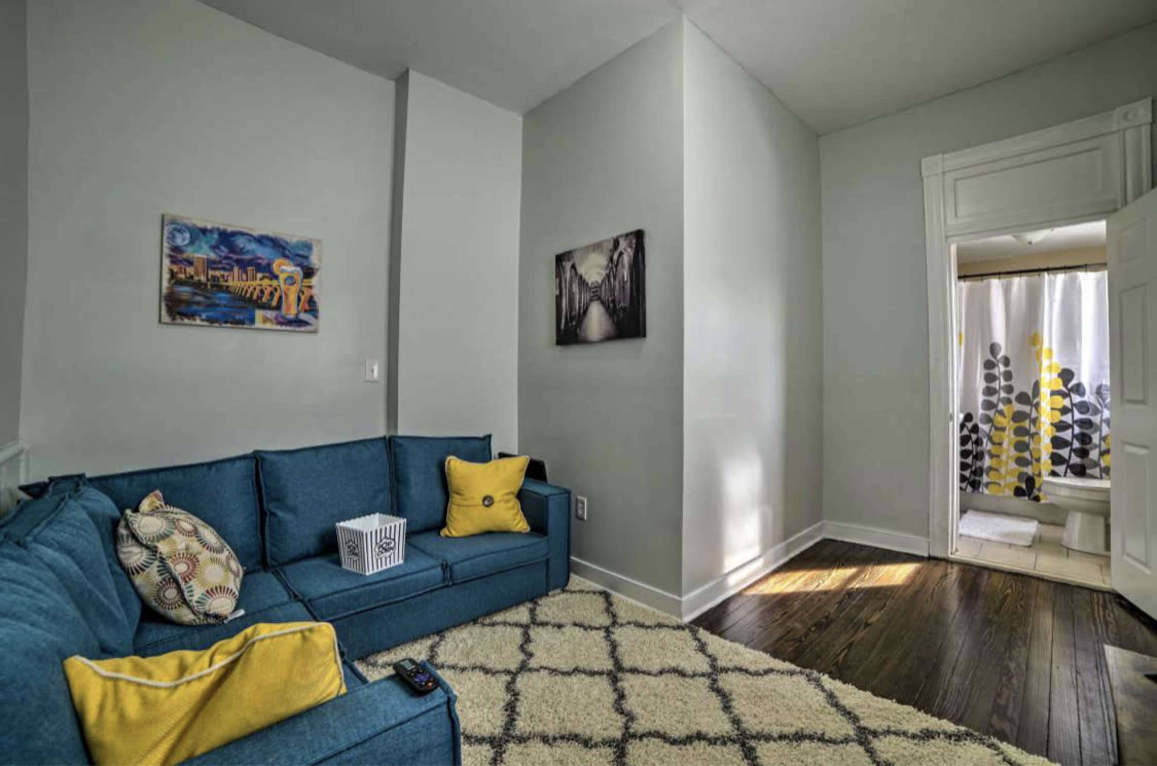 A bright living room features a blue couch with yellow accent pillows, a decorative rug, and artwork on the walls, leading to an adjoining bathroom area.