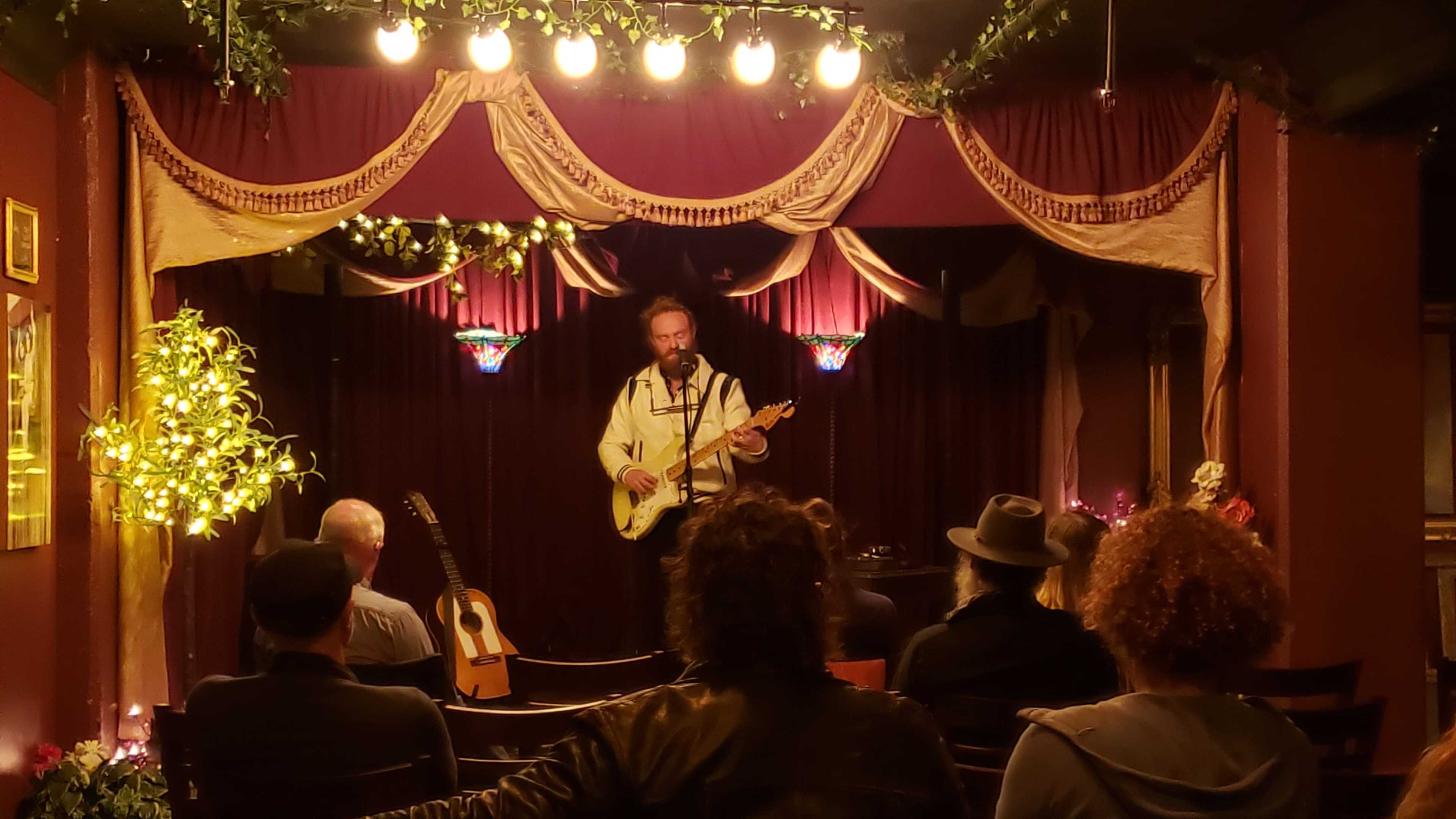 A musician performs on stage in a small venue with decorative lighting and an audience seated in front.