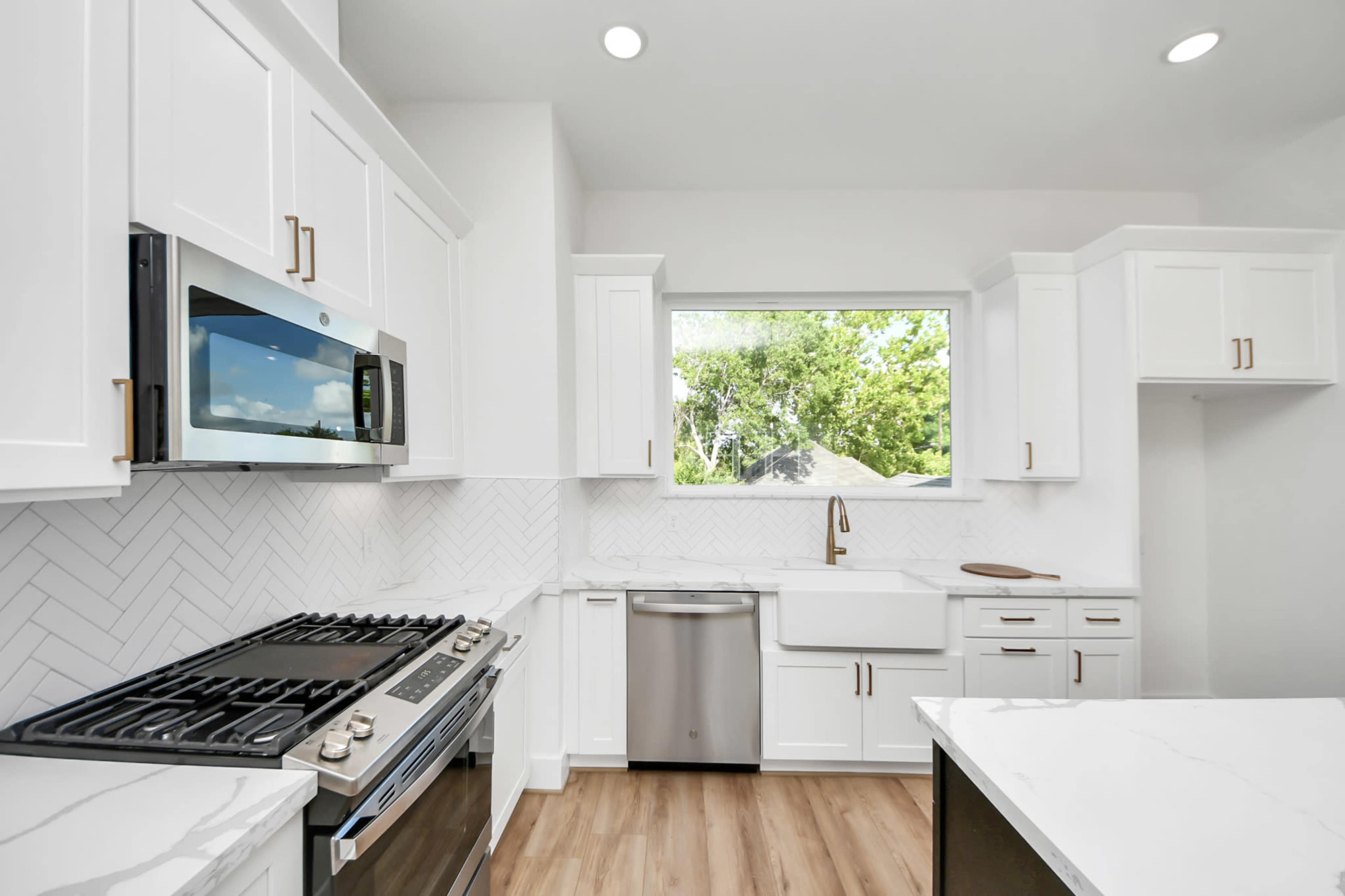 The image shows a modern kitchen featuring white cabinetry, a stainless steel microwave, a gas range, and a window overlooking greenery.
