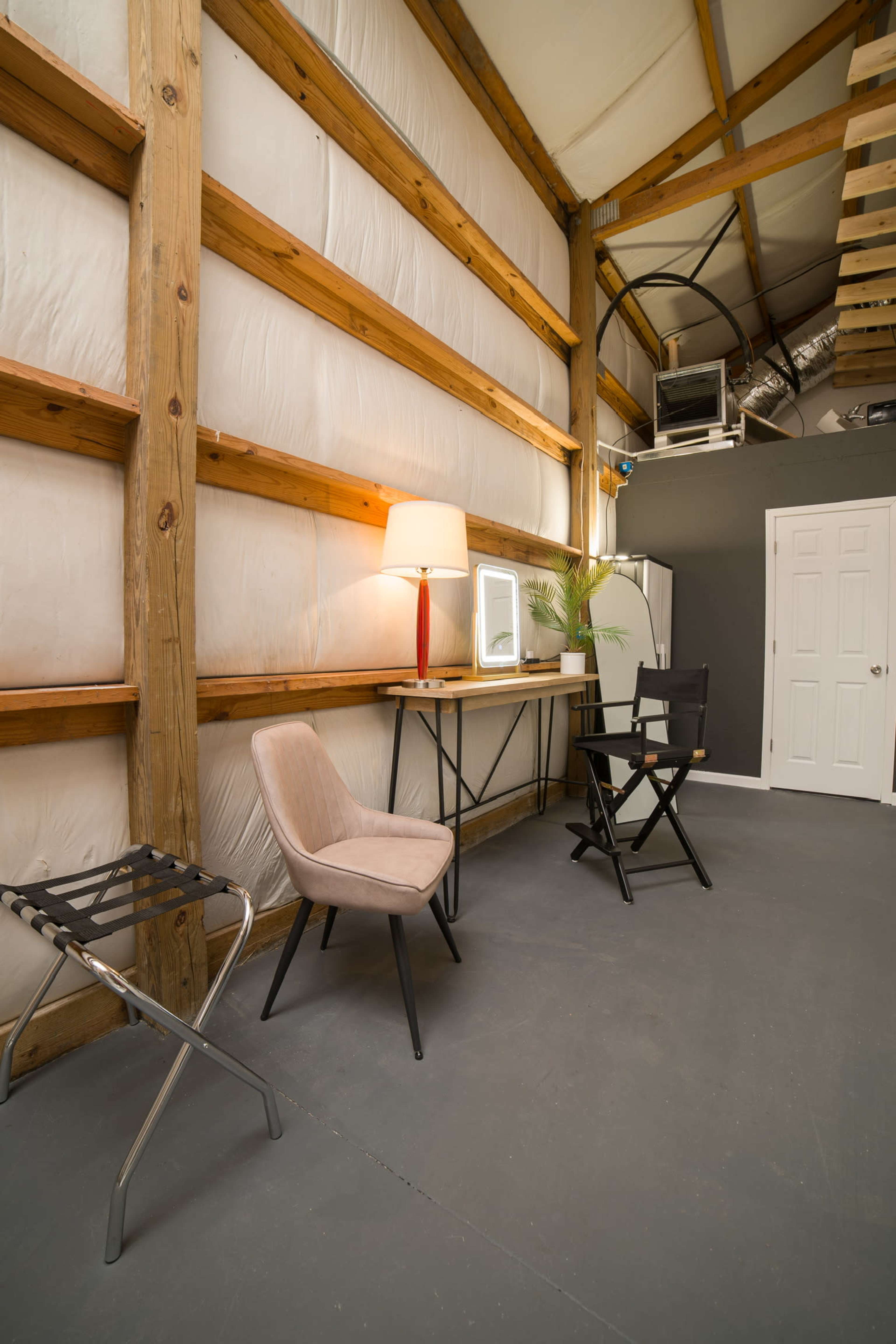 The image shows a spacious interior featuring wooden beams, a desk with a mirror, a lamp, and two chairs on a gray concrete floor.