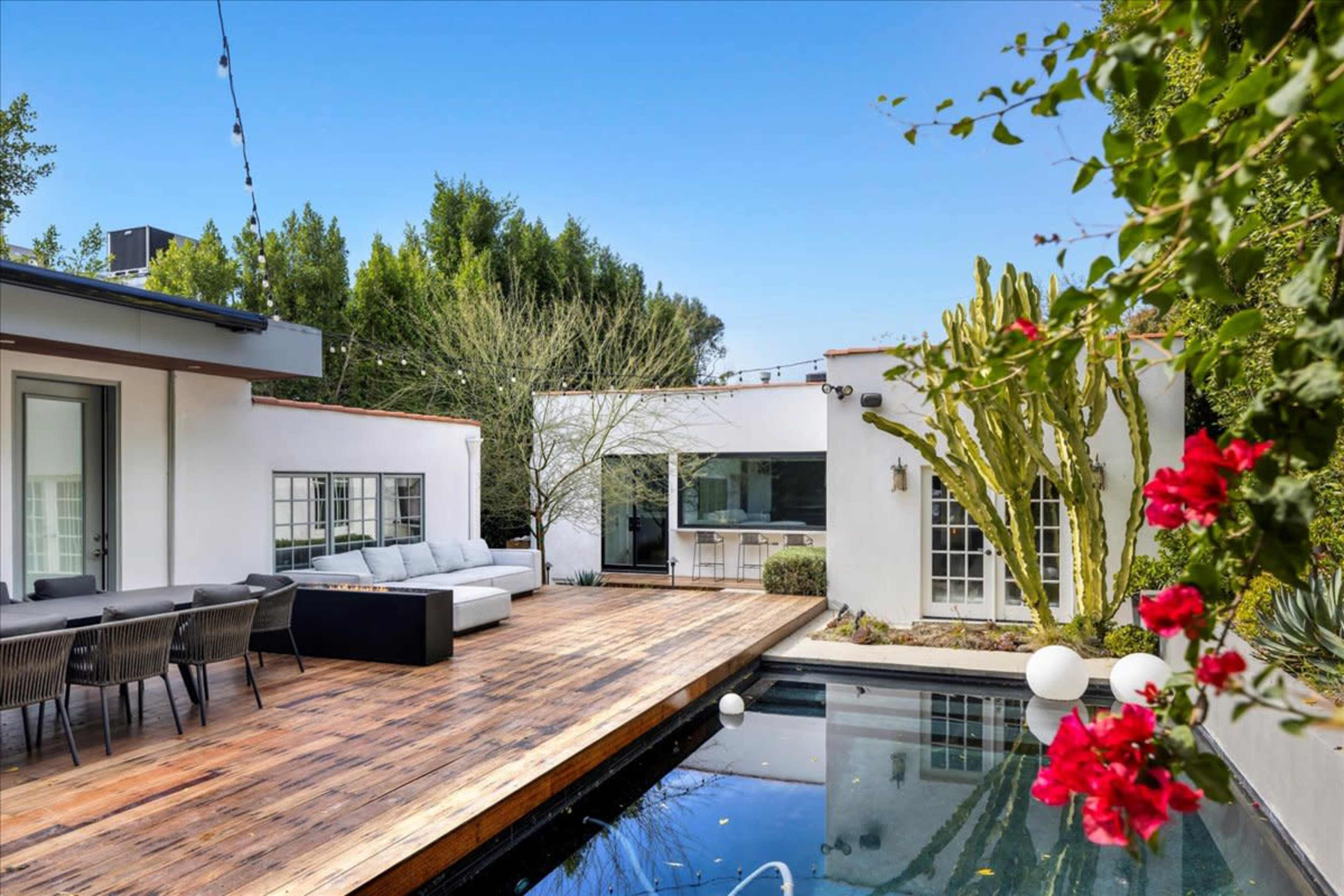 A modern outdoor space features a wooden deck with seating and a swimming pool surrounded by lush greenery and flowering plants.