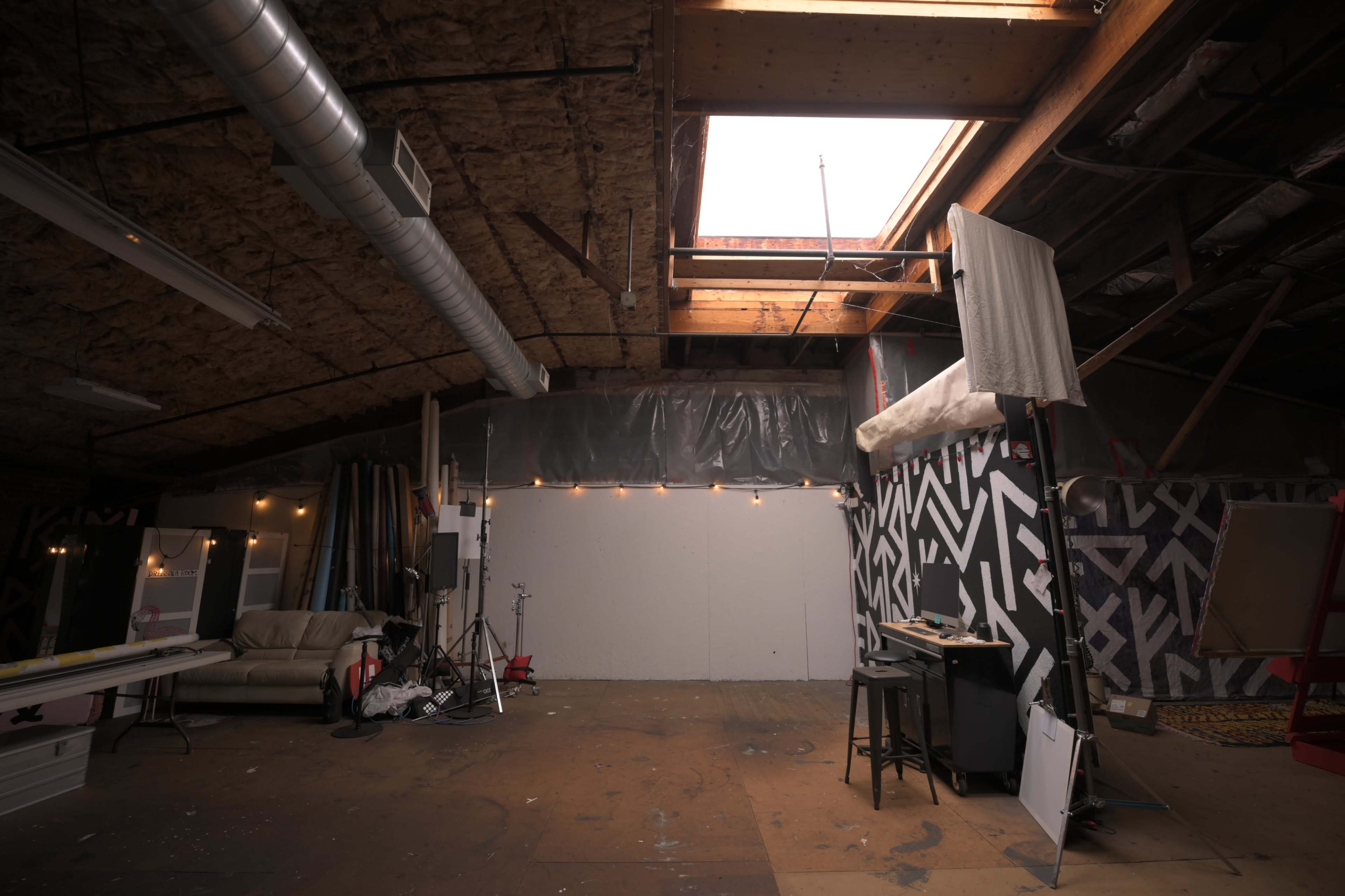 The image shows a spacious, unfinished interior of a studio with a skylight, various tools, furniture, and a contrasting patterned wall.