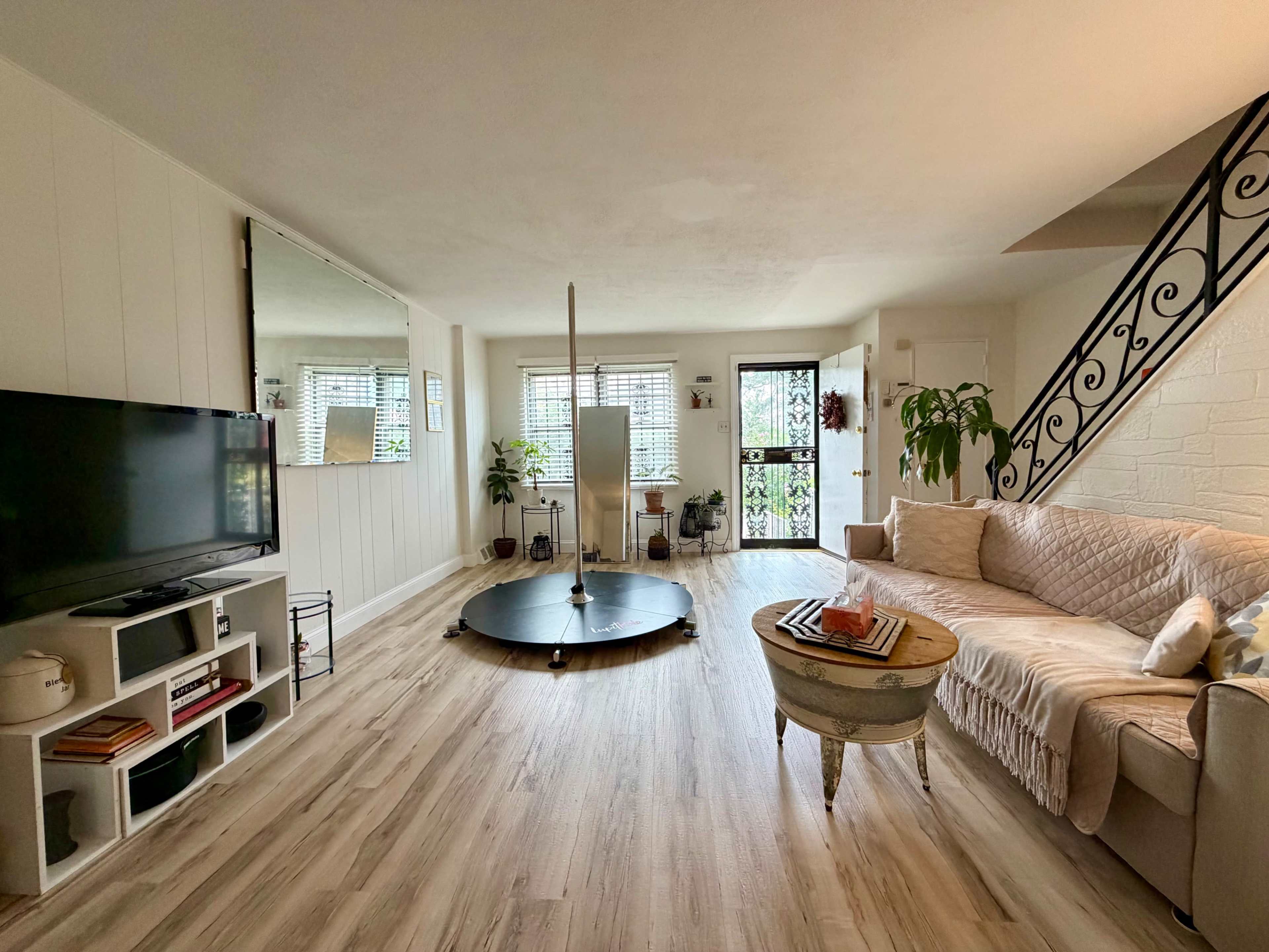 The image shows a spacious living room with a pole in the center, a couch, a television on a stand, and a staircase in the background, alongside large windows with plants.