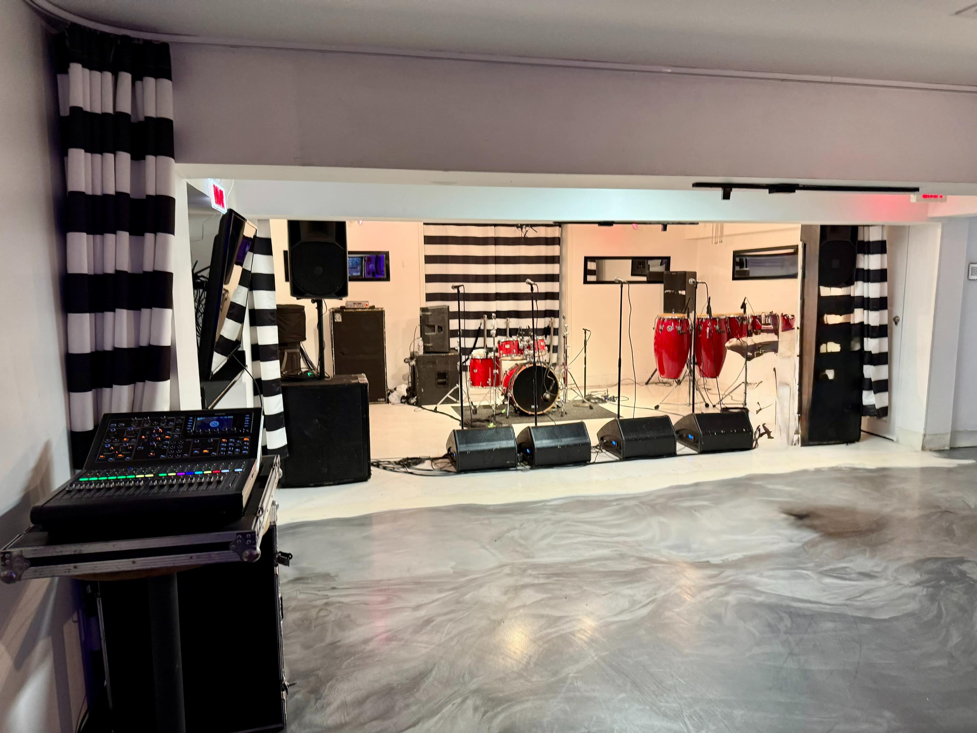 A performance space features a stage with drums and congas, flanked by sound equipment and decorated with black and white striped walls.