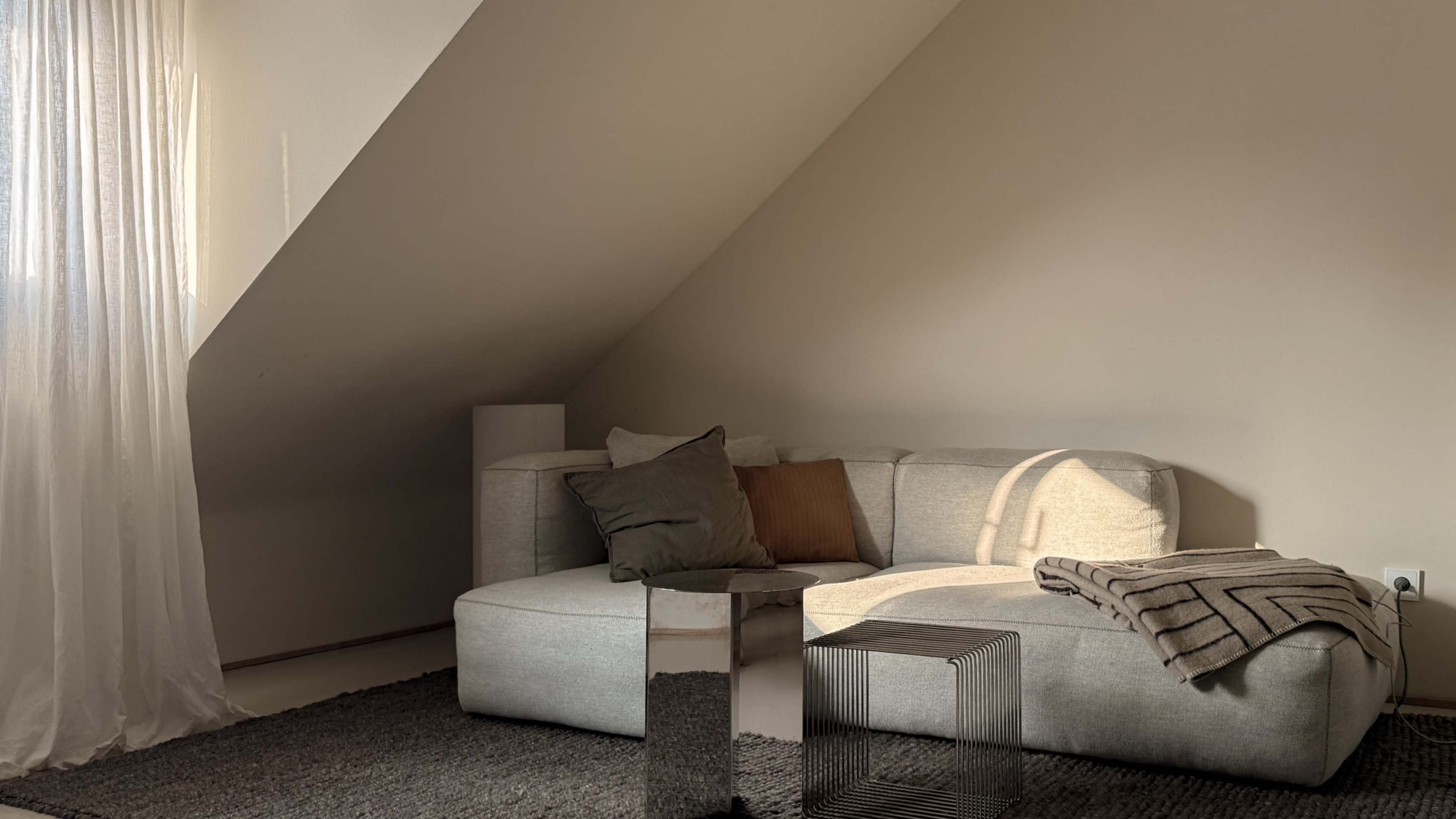The image shows a modern, minimalist living space with a light gray sectional sofa, a small metallic side table, and soft lighting casting shadows on the walls.