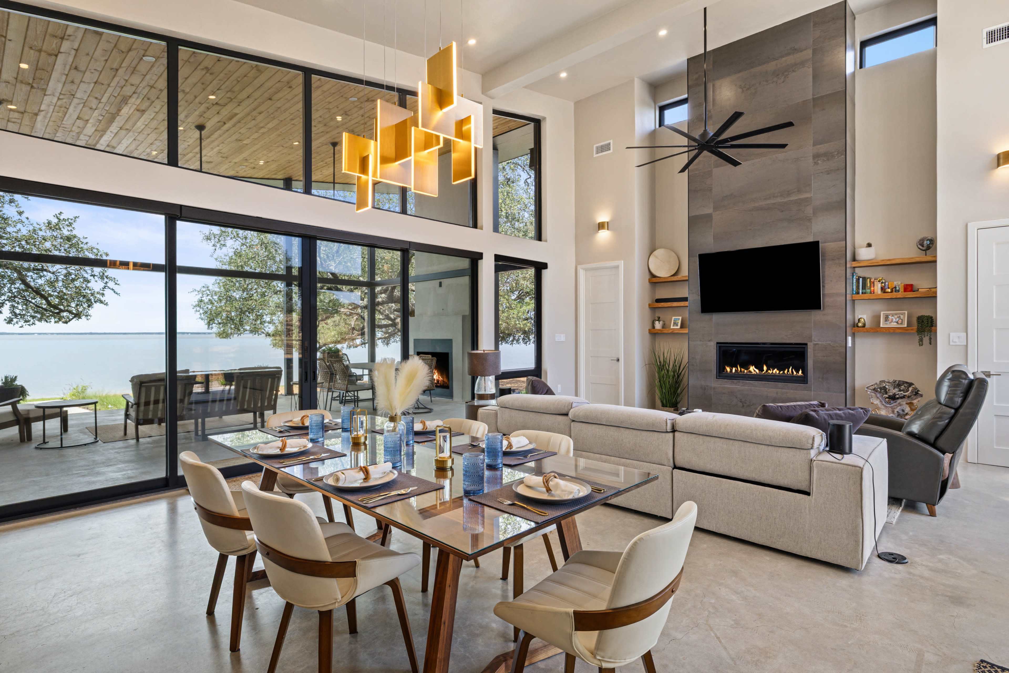 A modern dining area features a glass table set for a meal, with a spacious living room and large windows overlooking a body of water.