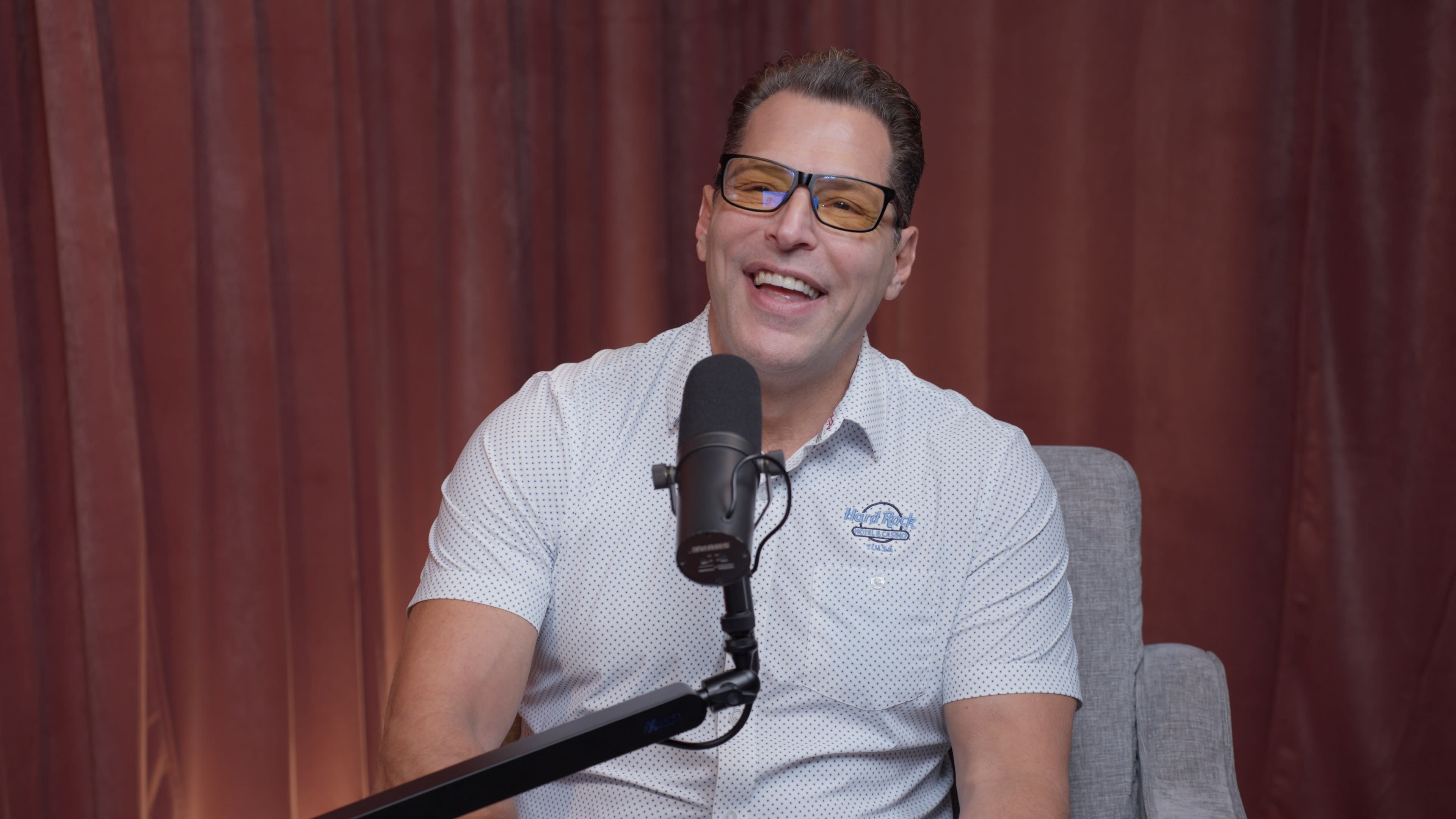 A man wearing glasses smiles while sitting in front of a microphone in a studio setting.